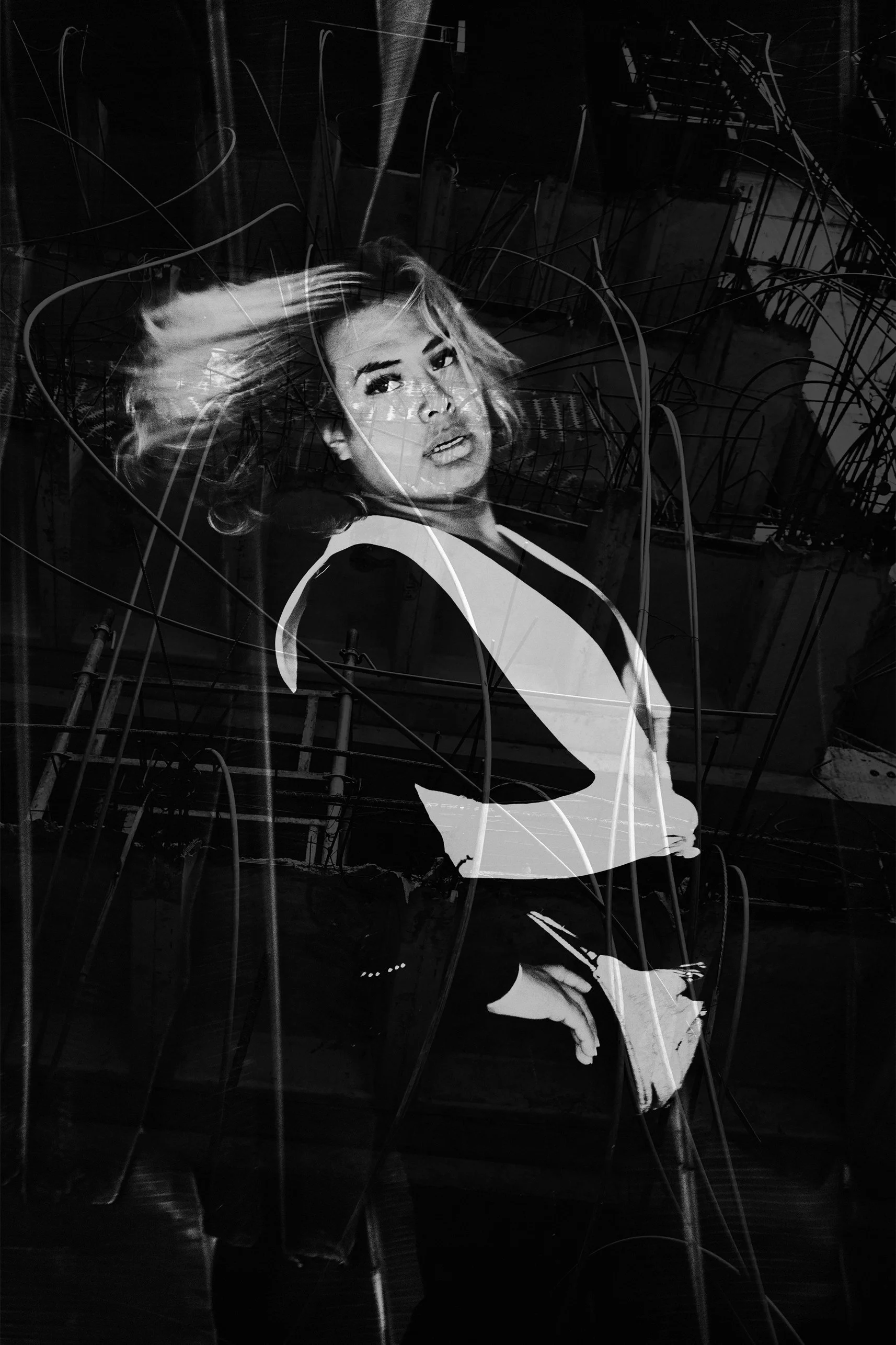 Retrato en negro y blanco de una mujer con cabello rubio suelto, usando una blusa blanca y pantalones oscuros, posando con la mano en la cadera, con un fondo de líneas y estructuras abstractas.