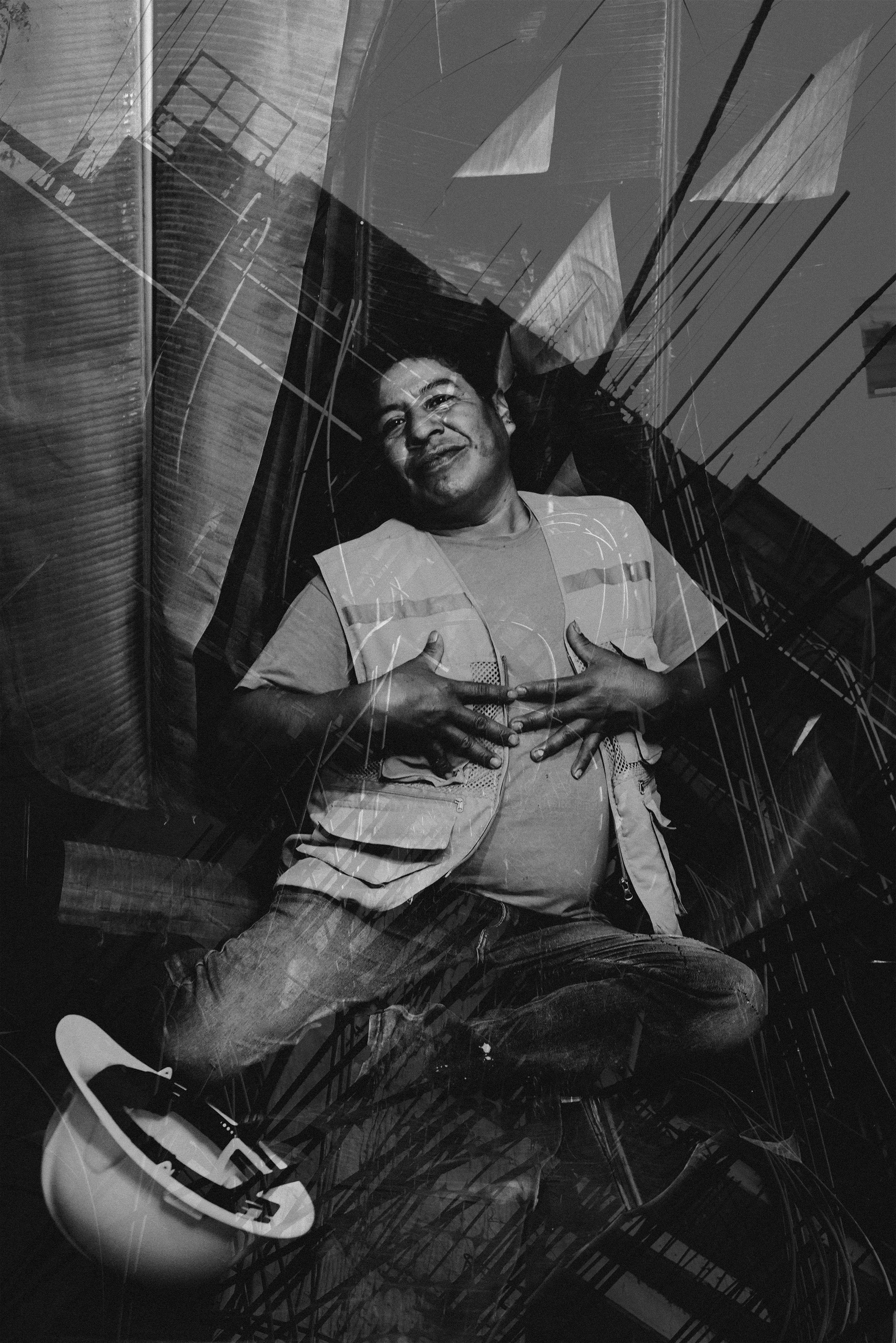 Hombre sonriente acostado sobre la acera, con ropa casual y casco de motocicleta a su lado, reflejo de edificios y líneas eléctricas en el vidrio