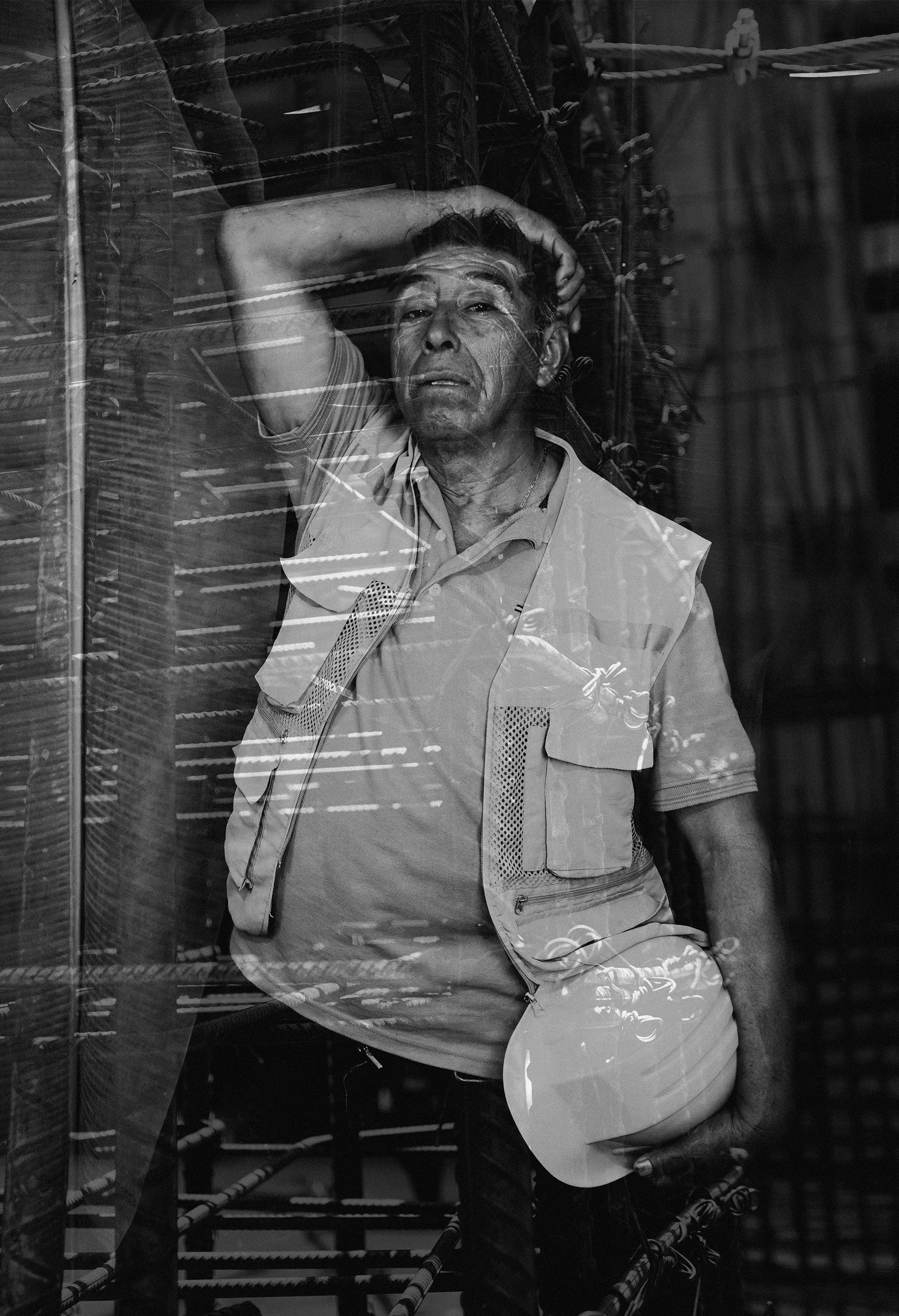 Hombre mayor con expresión de cansancio o estrés, apoyado contra una superficie, con un brazo detrás de la cabeza, visto a través de un cristal reflejando líneas y objetos del entorno.