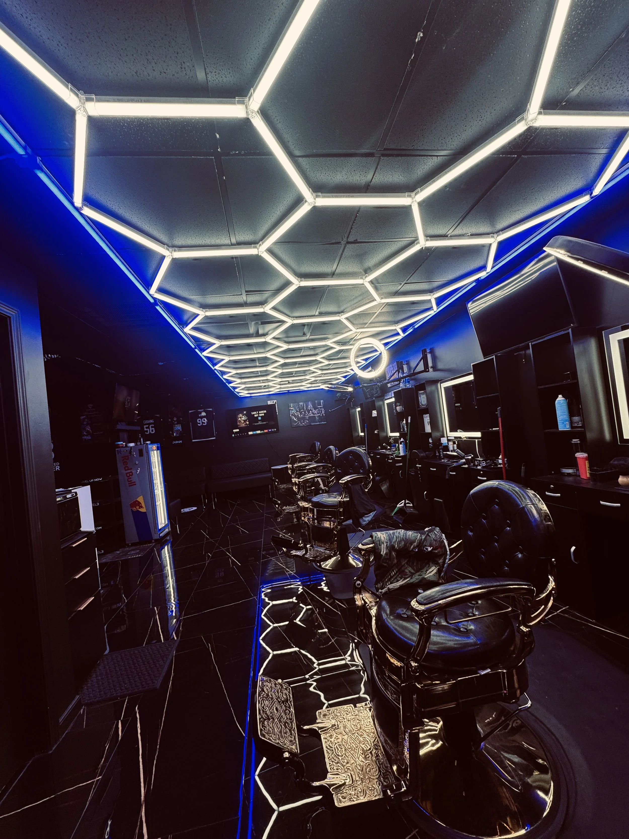 A modern barbershop or salon with black walls, illuminated hexagon-shaped light fixtures on the ceiling, and black furniture including barber chairs. There are mirrors with white lighting around them, and a small refrigerator with a Red Bull logo. The floor is black with reflective tiles.