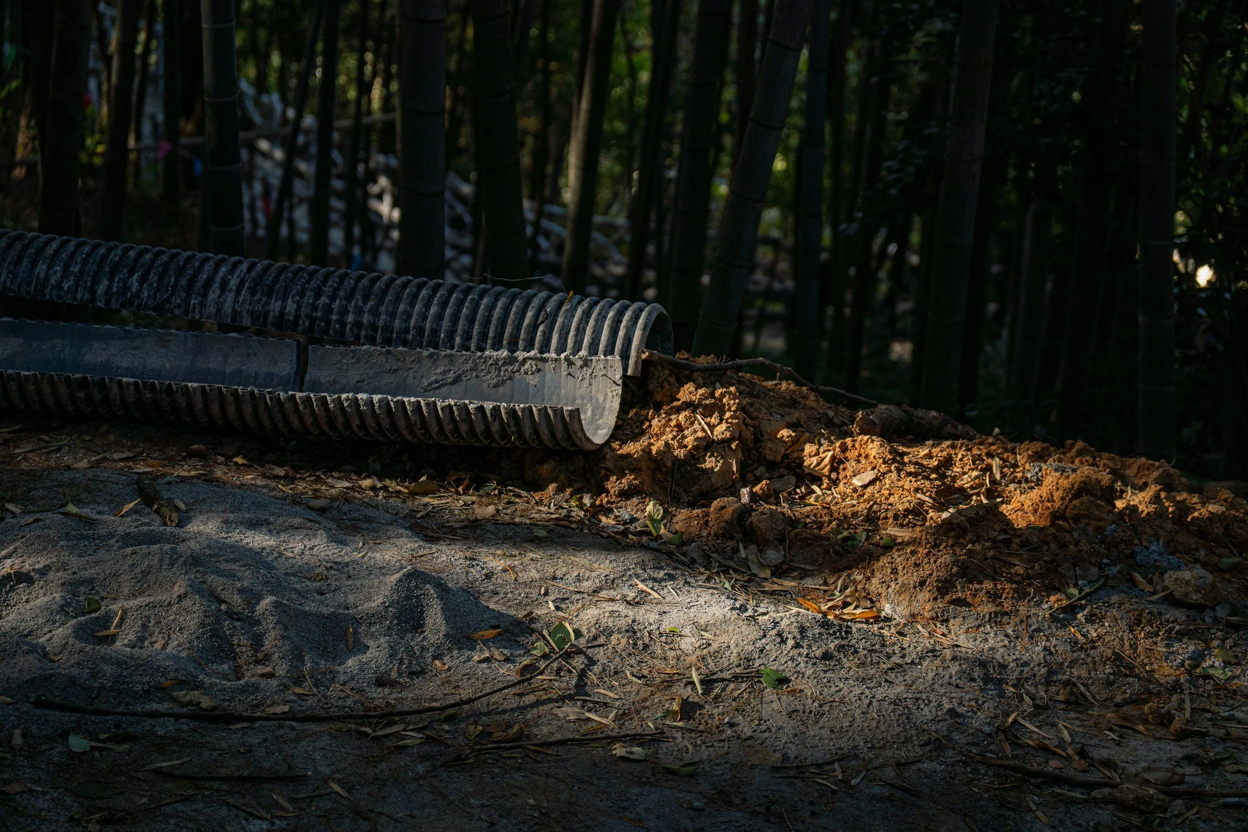 Close-up of a pipe laying in dirt with soil and small debris in front of dark bamboo forest.