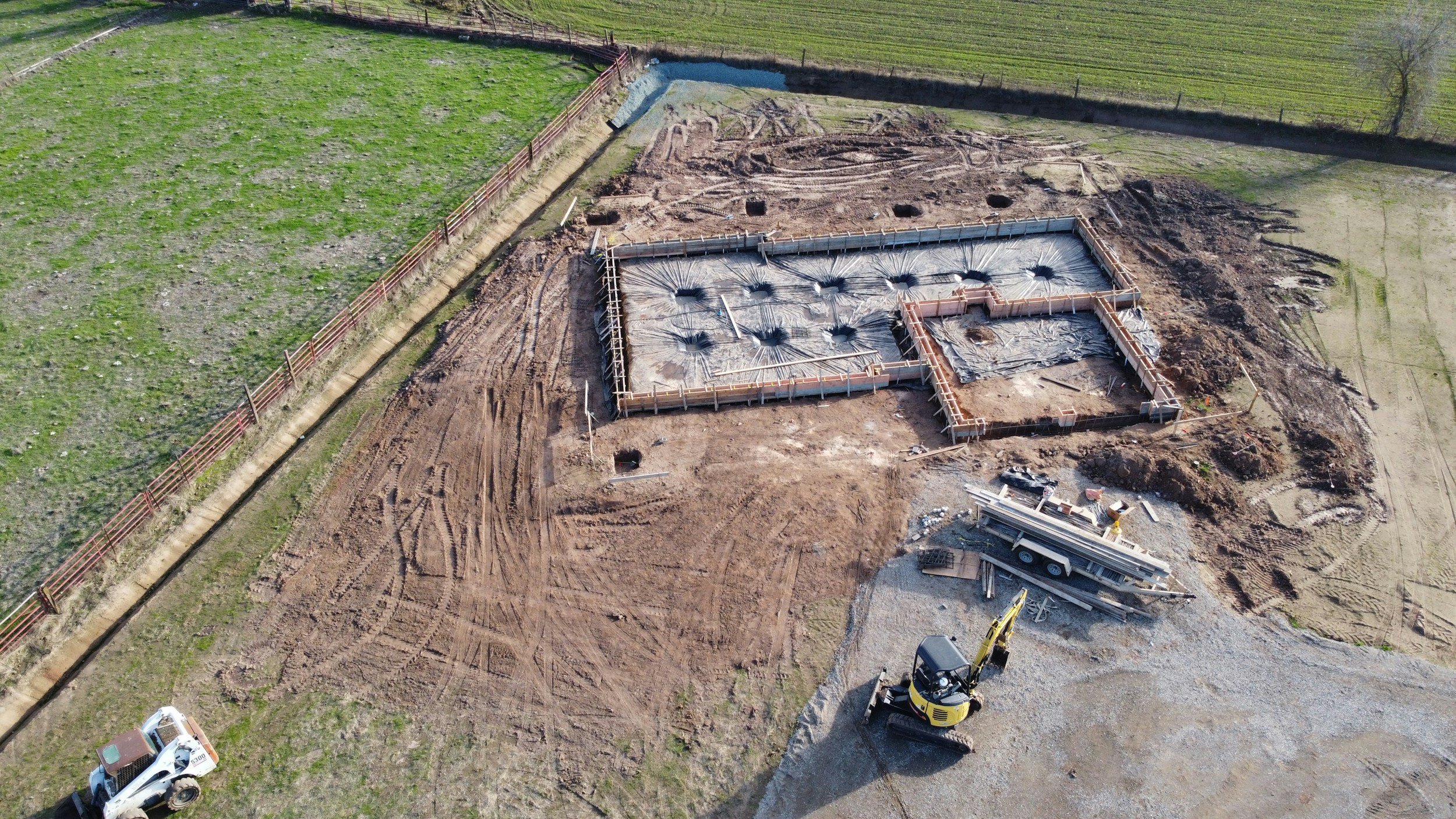 Aerial view of a construction site with foundation work in progress, concrete formwork, and construction vehicles, surrounded by green fields.