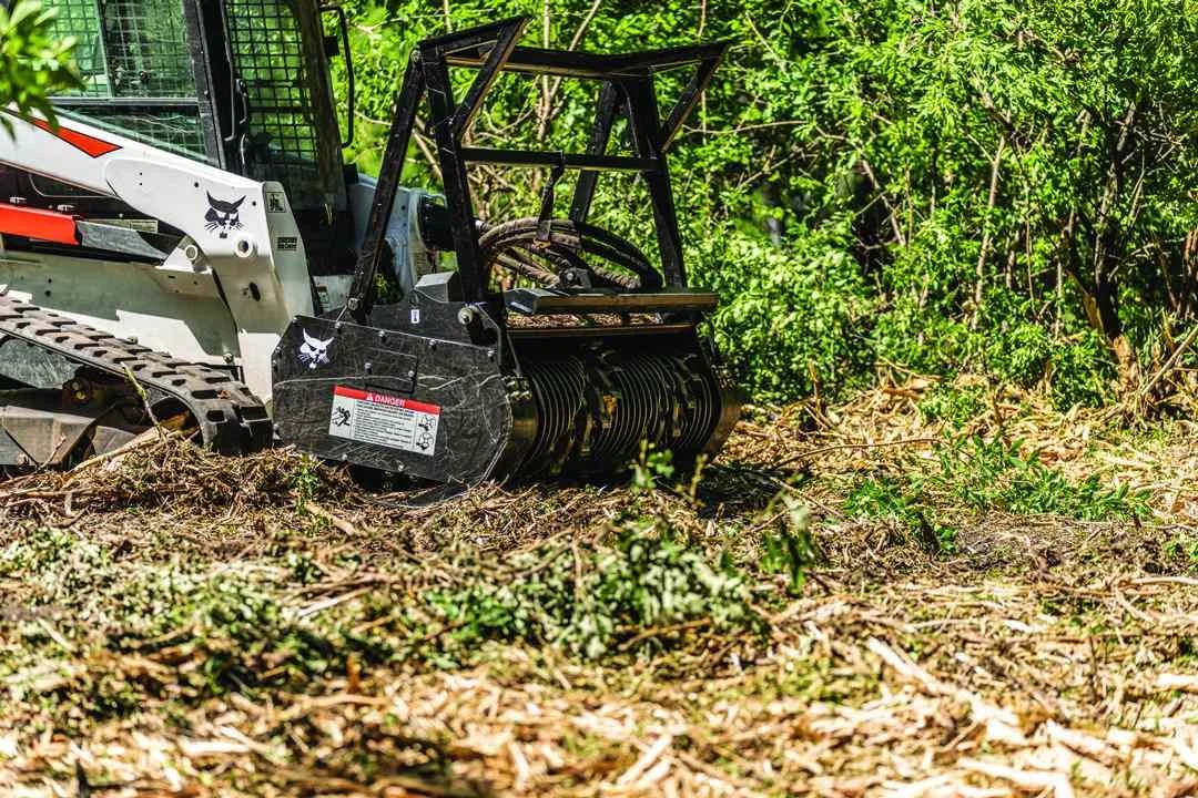 Forestry mulcher attached to a compact track loader in a wooded area, clearing vegetation.