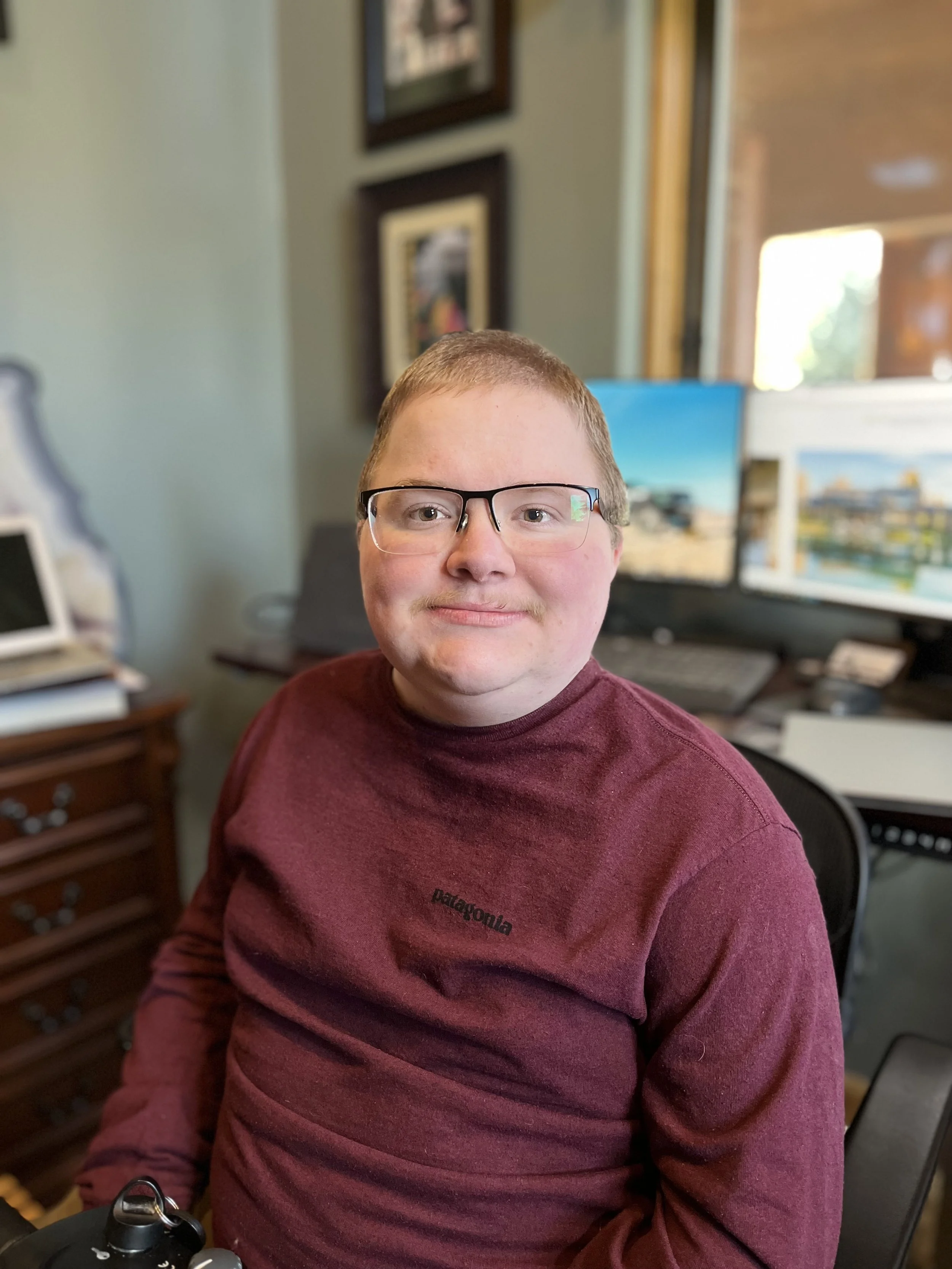 Person wearing glasses and a maroon sweater sitting in a home office with a desk, computer, and framed pictures in the background.