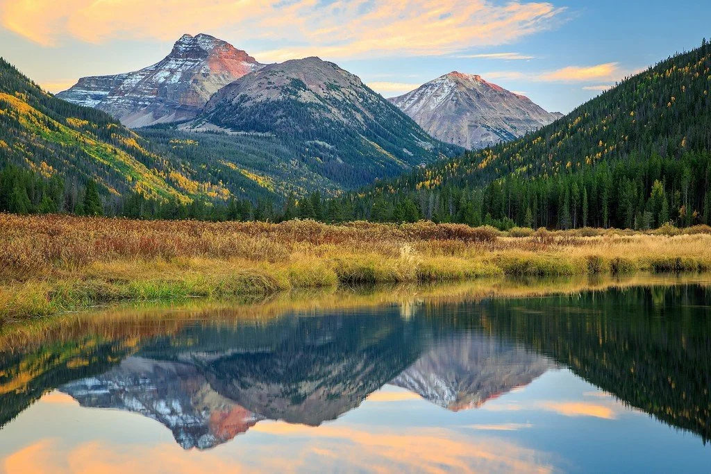 Scenic view of a lake reflecting surrounding mountains with snow-capped peaks, green forests, and a colorful sky during sunset or sunrise.