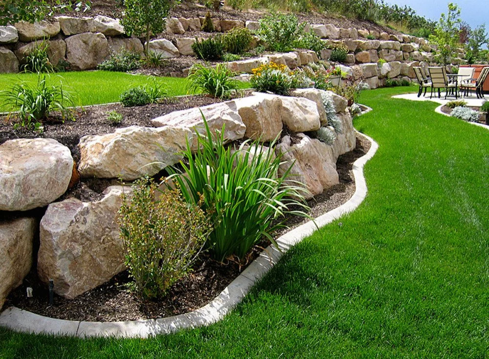 Terraced backyard with stone retaining walls and vibrant green lawn, featuring landscaped plants and a patio area with chairs.