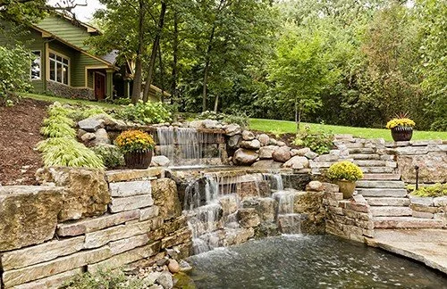 Landscape featuring a small waterfall cascading over rocks into a pond, surrounded by stone masonry and lush greenery. A house is partially visible in the background, with planters displaying blooming flowers alongside the waterfall.