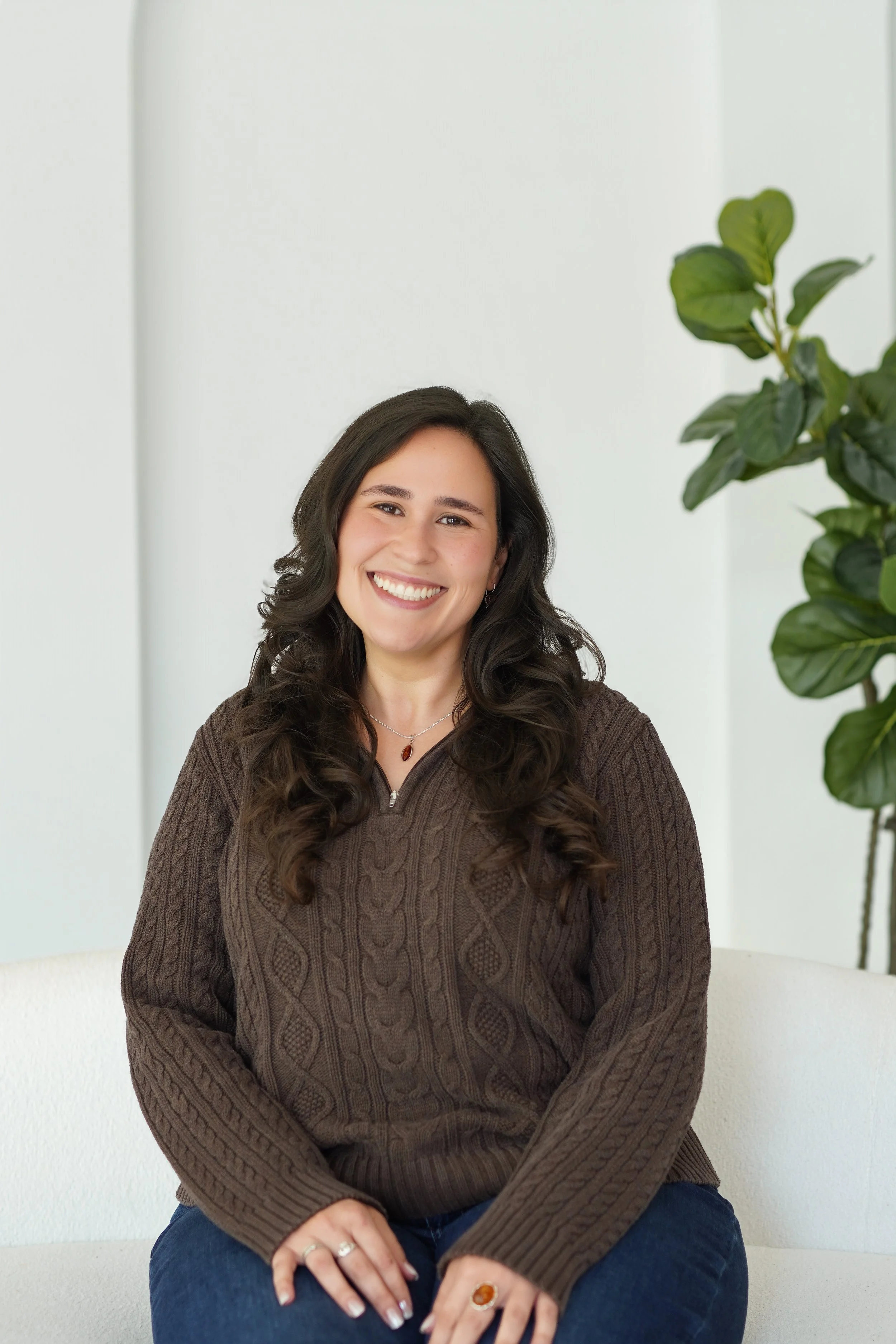 A woman with long dark wavy hair and light skin, smiling, wearing a brown cable-knit sweater, a necklace with a red pendant, and rings on her fingers, sitting in front of a white wall and a green leafy plant.