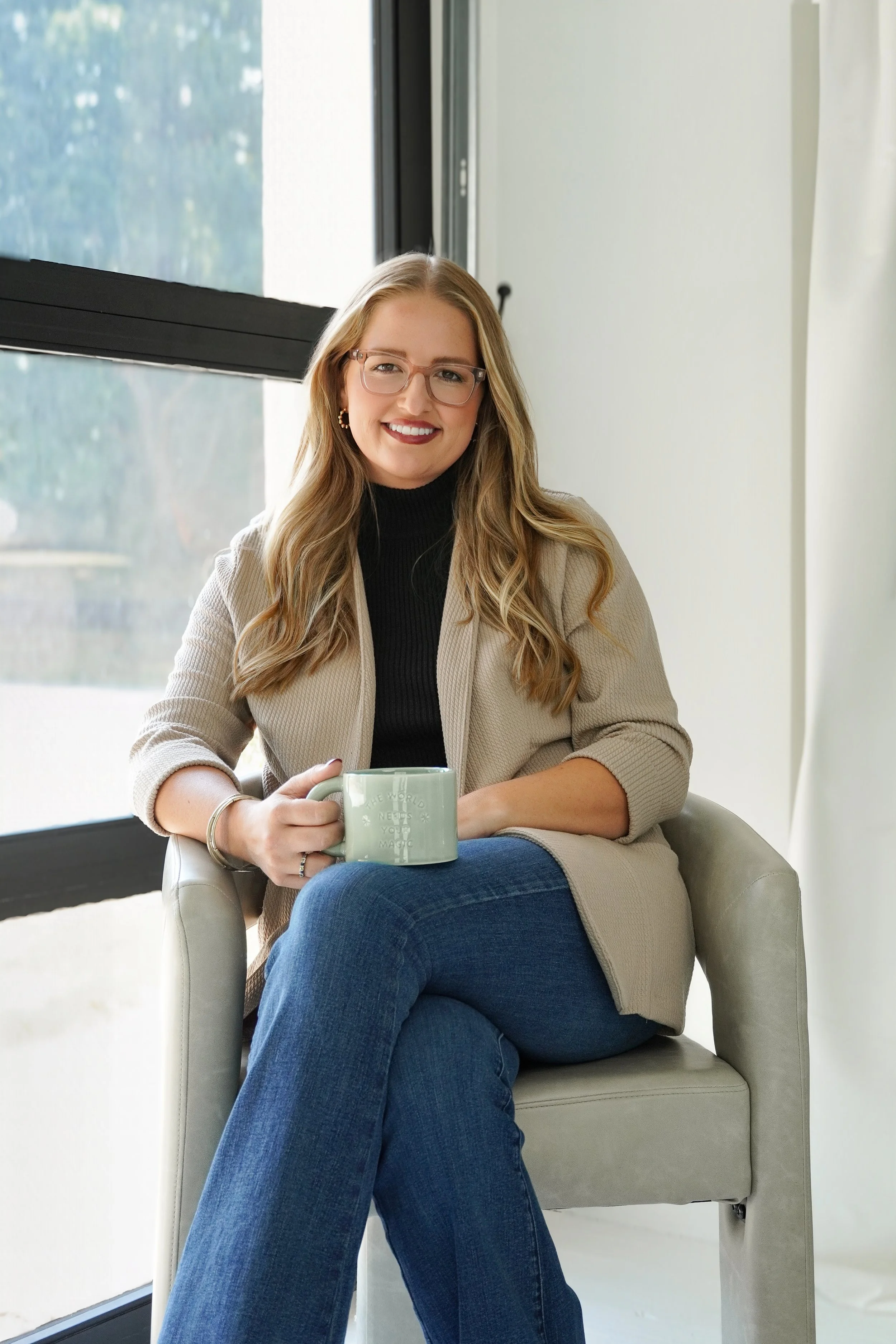 Young woman with long wavy blonde hair, glasses, and earrings, sitting in a light-colored armchair near a large window. She is wearing a black turtleneck sweater and a beige blazer, holding a mug, and smiling at the camera.