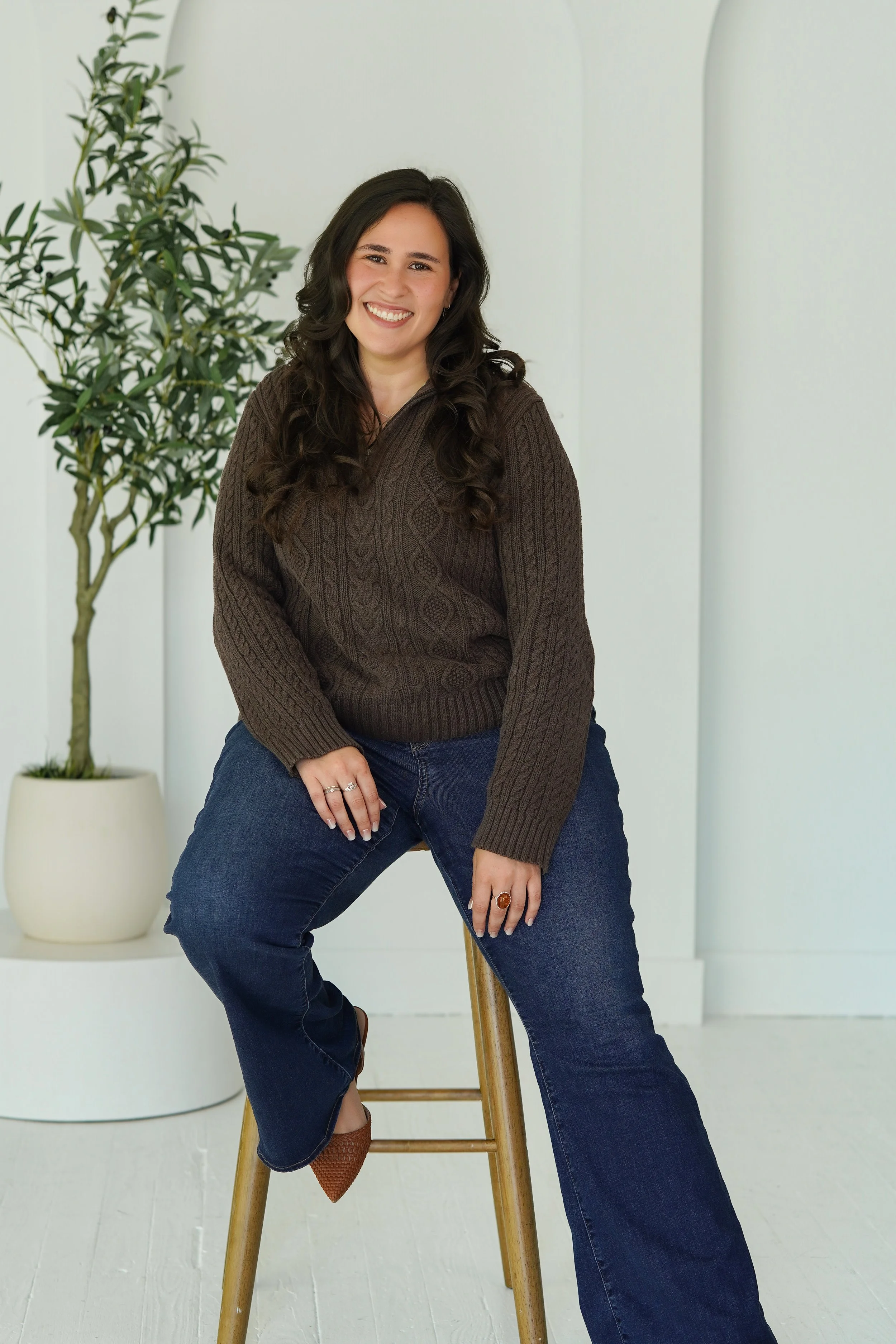 A woman with long dark curly hair smiling while sitting on a wooden stool, wearing a brown sweater and blue jeans. She poses with her left hand resting on her thigh and her right hand on her knee, in front of a white wall with a potted plant nearby.