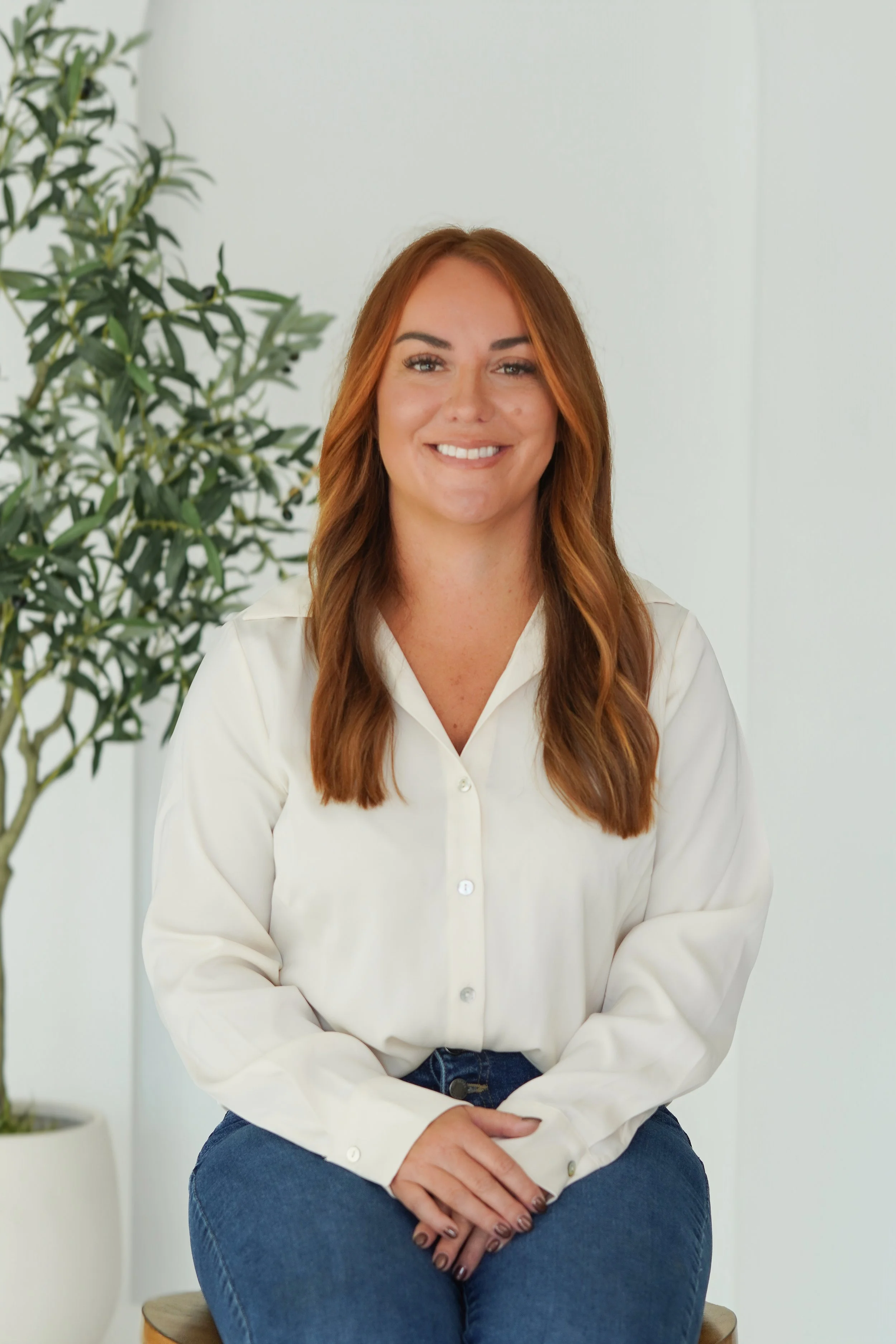 Young woman with long red hair, wearing a white button-up shirt and blue jeans, sitting cross-legged on a small stool in front of a white wall with a green potted plant in the background.