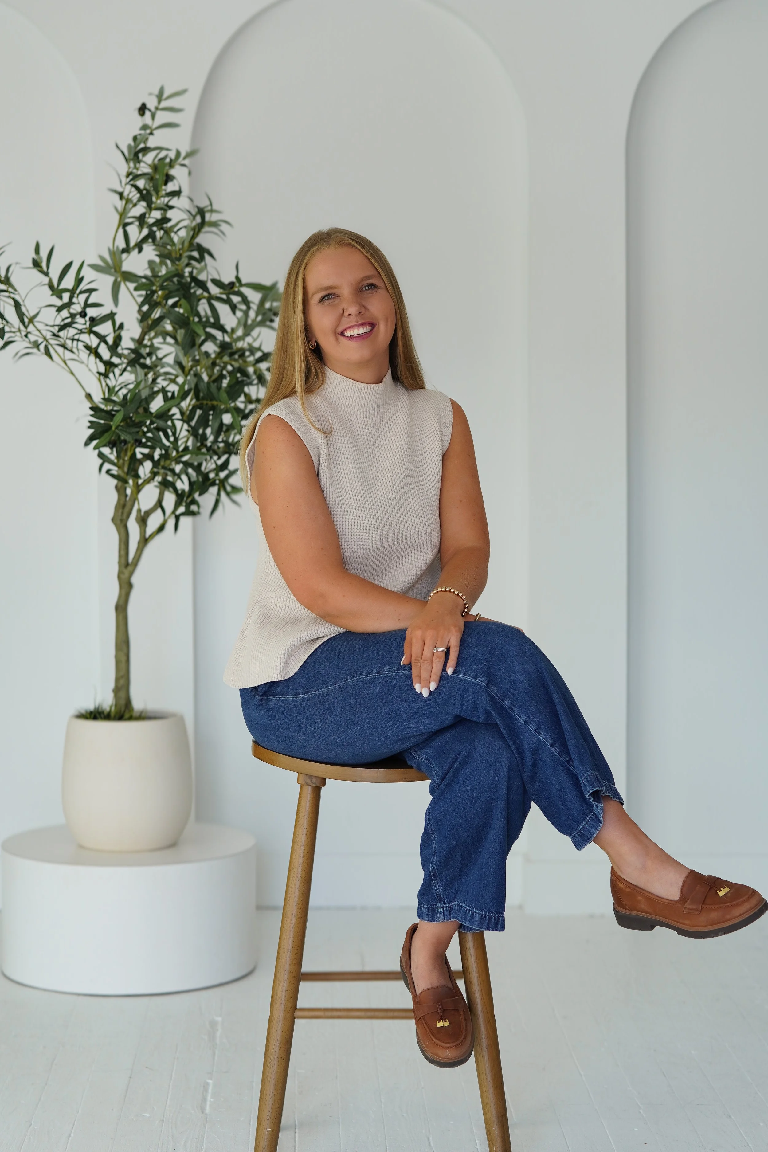 A woman with long blonde hair sitting on a wooden stool, smiling, arms crossed, wearing a sleeveless white top, blue jeans, and brown loafers, with a potted plant with green leaves in the background.