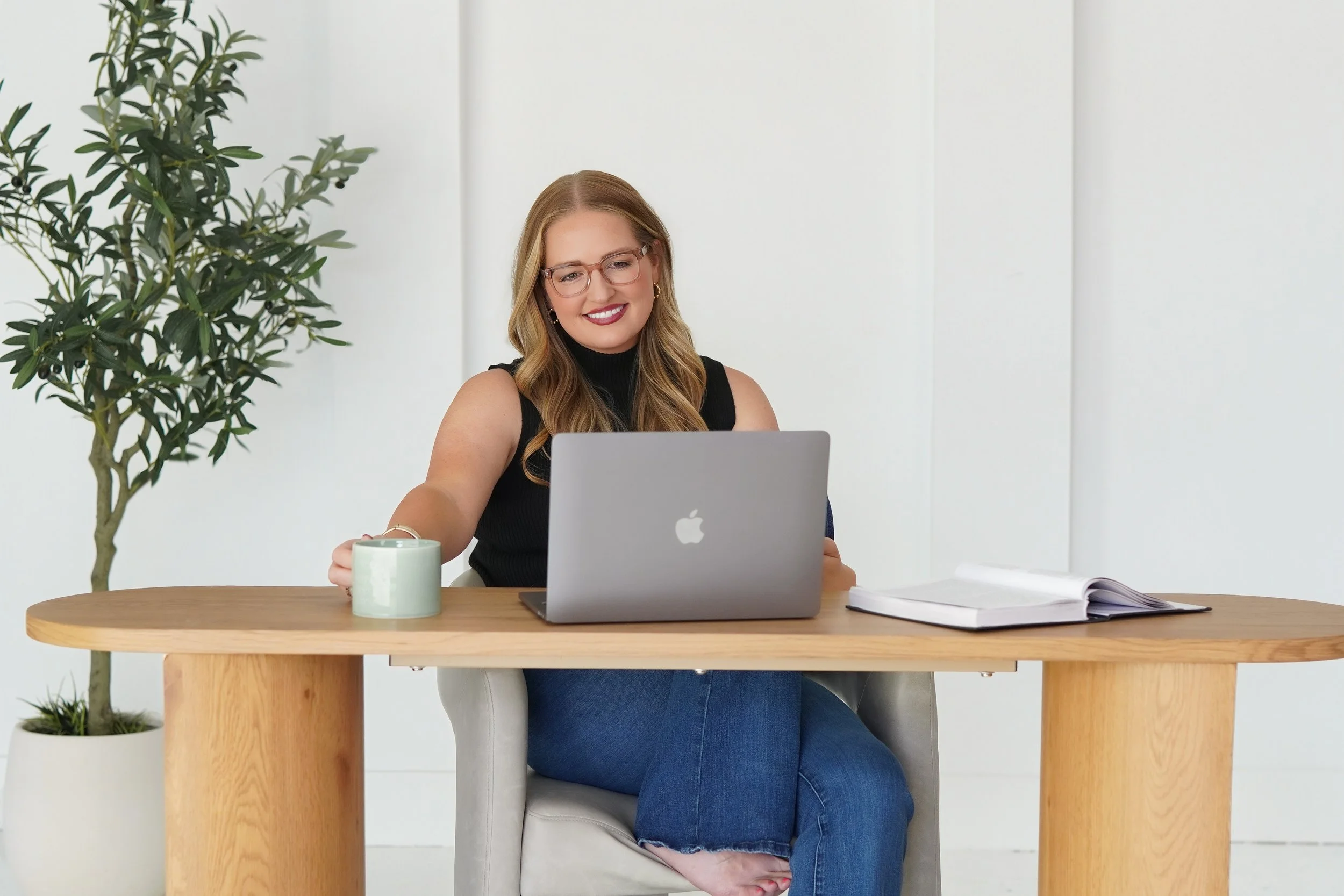 A woman with blonde hair, glasses, and a black sleeveless top sits at a wooden table in front of a silver MacBook laptop. She is smiling and holding a green mug. There is a large plant on her left and open books or notebooks on her right against a white background.
