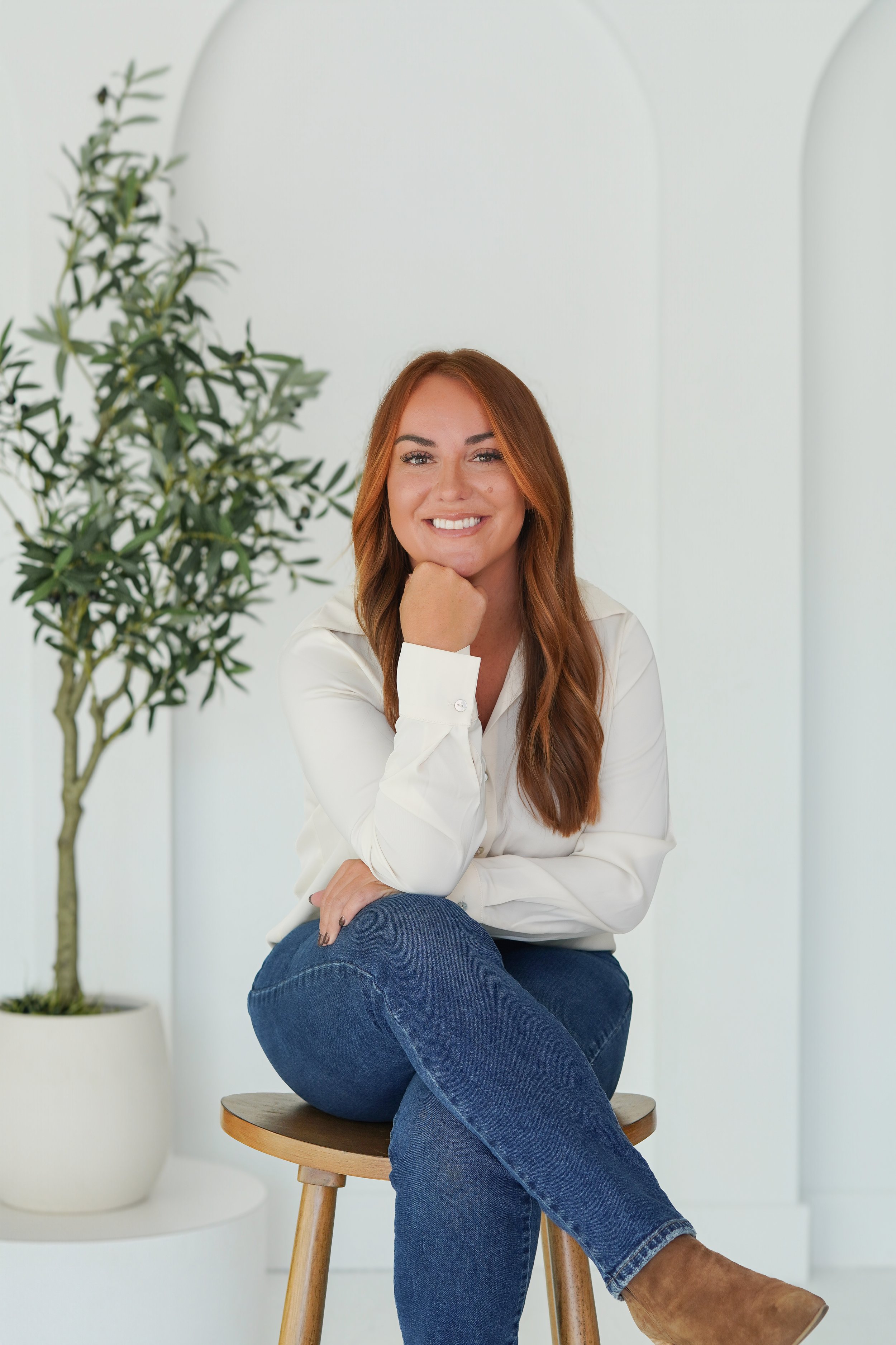 A woman with long red hair smiling while sitting on a wooden stool in front of a white background and green plant, wearing a white shirt, blue jeans, and brown boots.