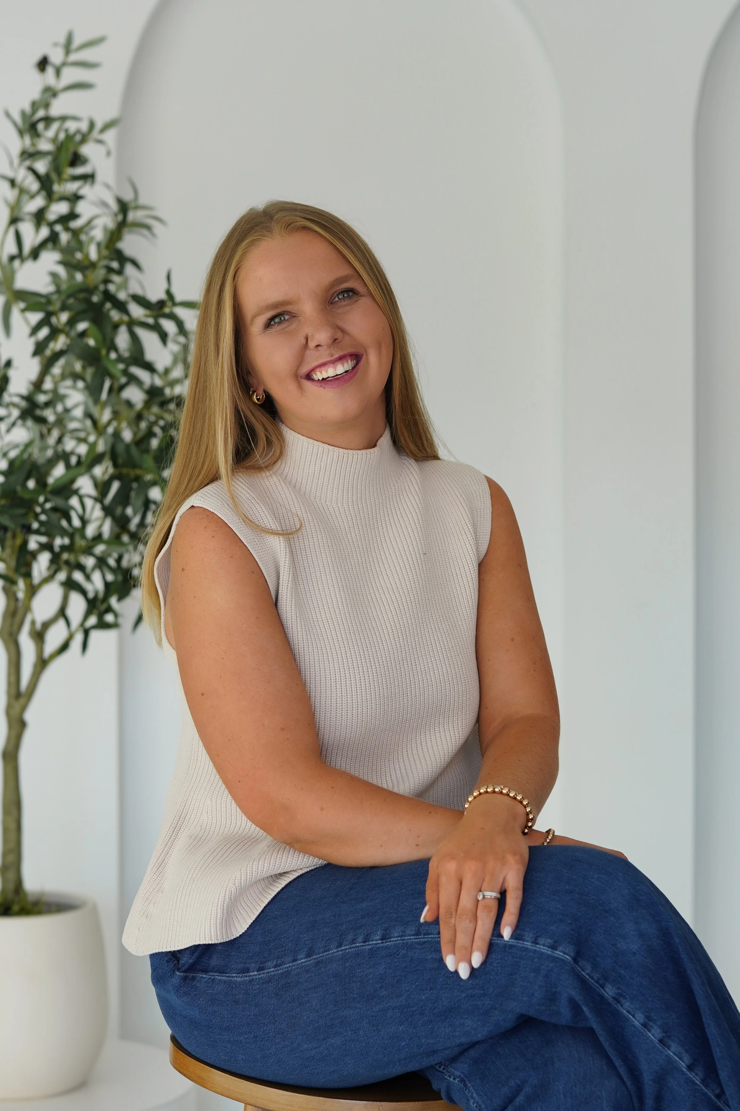 A smiling woman with blonde hair, wearing a sleeveless white top, sitting on a round stool with her legs crossed, in a bright room with white walls and a green plant in the background.