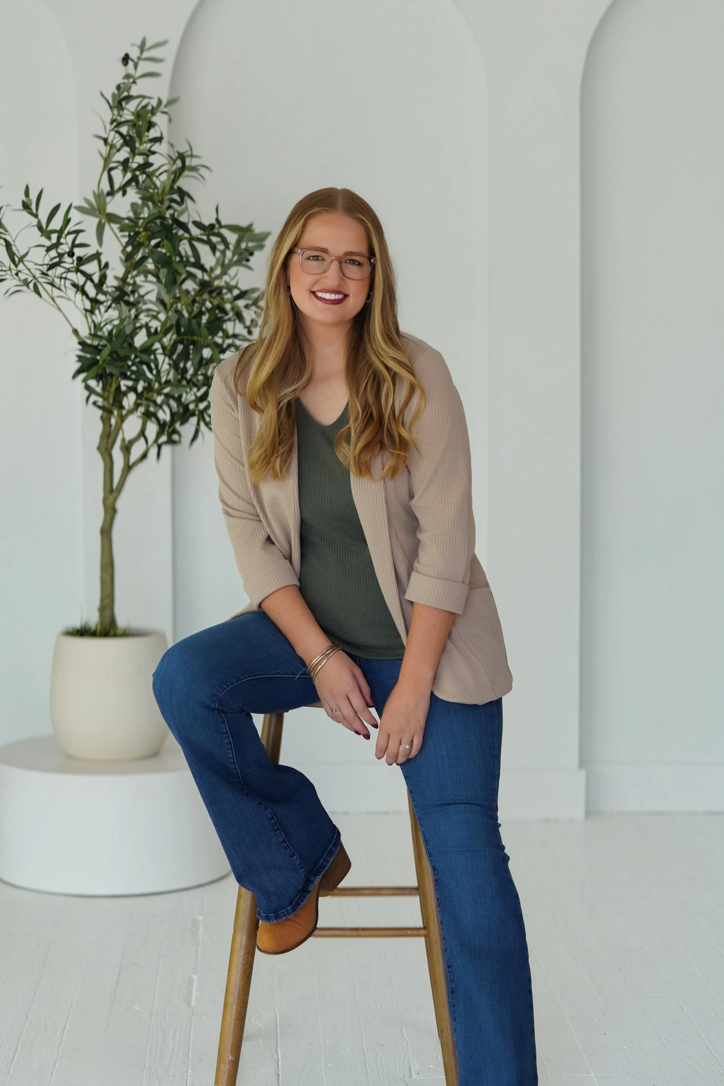 A woman with long blonde hair, wearing glasses, a beige blazer, a green top, and blue jeans, sitting on a wooden stool in a white room with minimal decor. Behind her is a potted plant with green leaves.