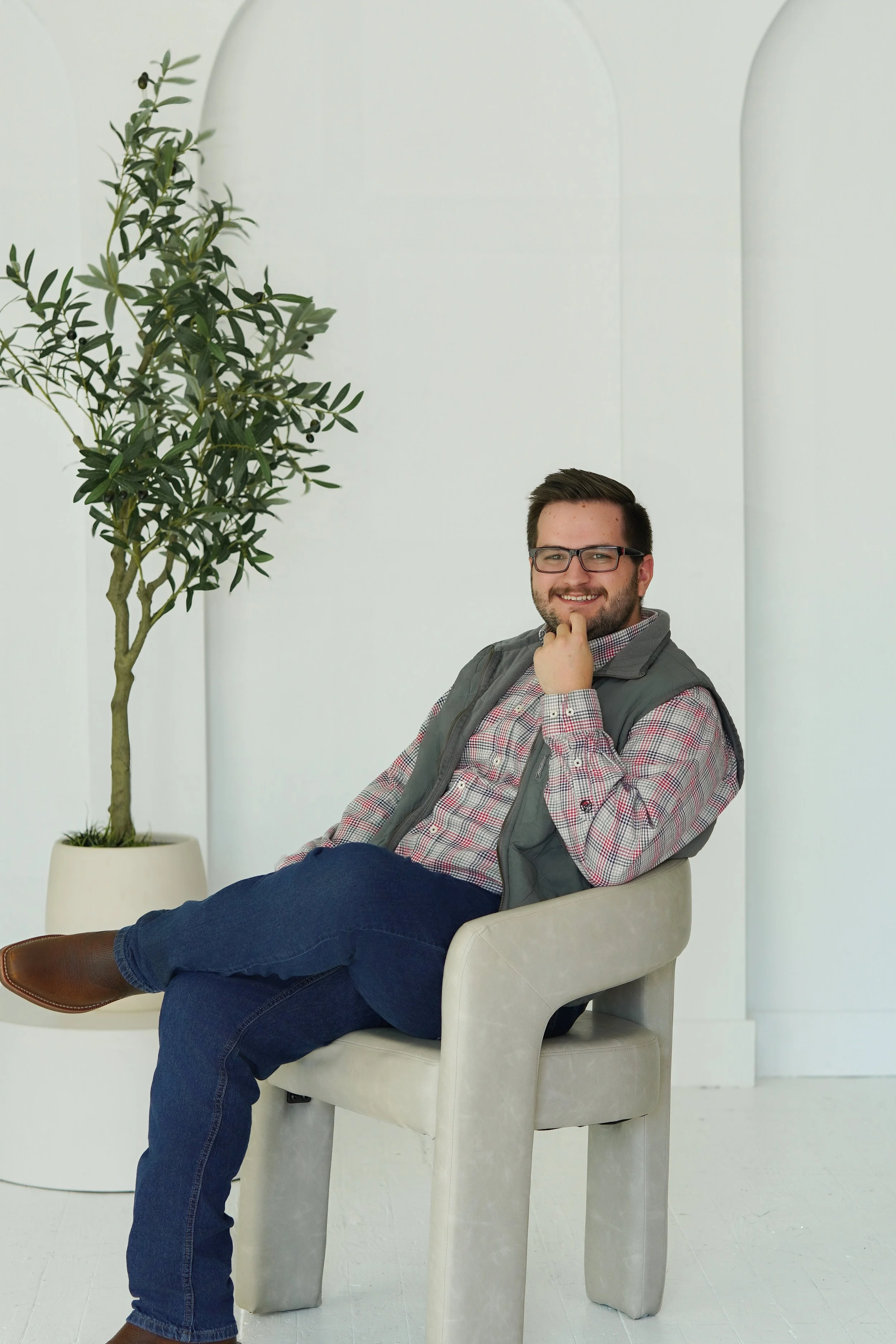 A man with glasses sitting in a beige armchair, smiling, with his legs crossed. A large potted tree with green leaves is positioned behind him to the left. The background features a minimalist white wall with arch shapes.