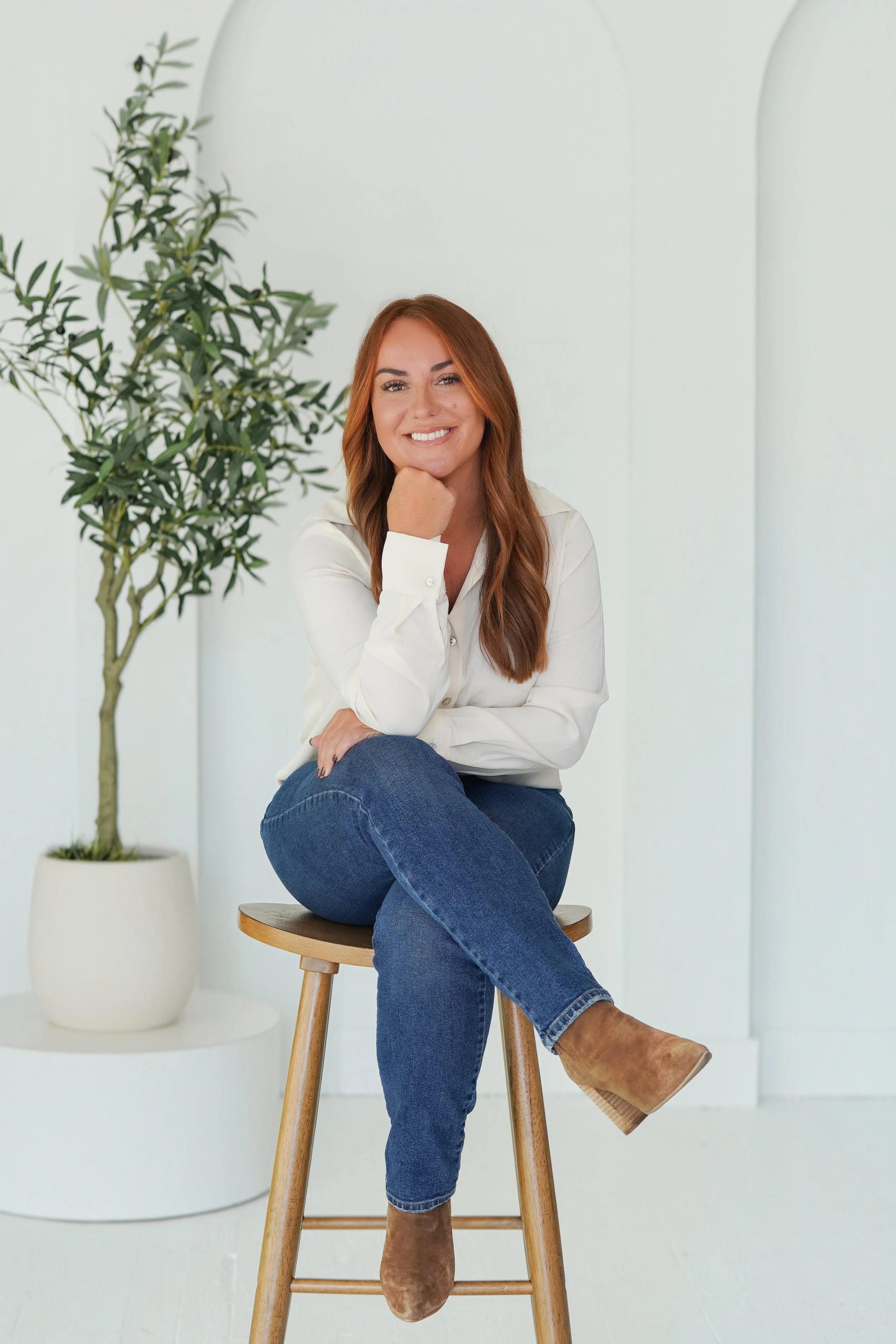 A woman with long auburn hair, wearing a white blouse, blue jeans, and tan ankle boots, sitting on a wooden stool in front of a white background with a potted plant nearby.
