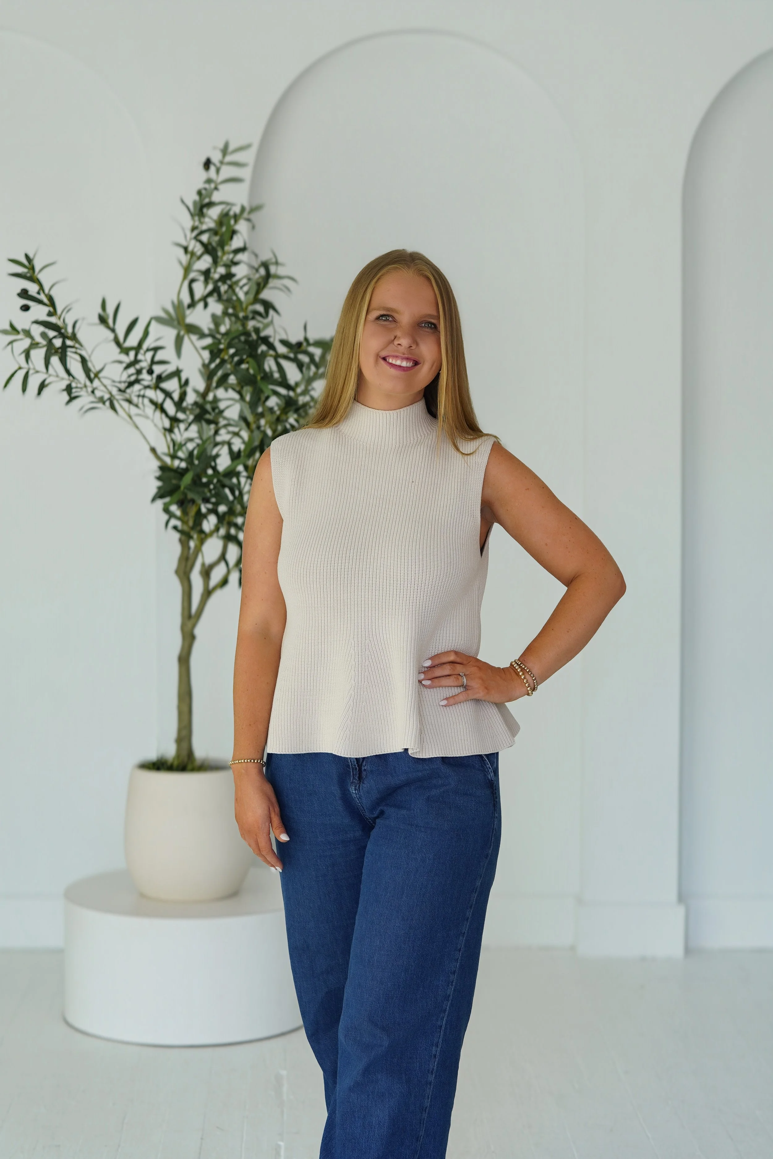 A woman with blonde hair wearing a sleeveless white top and blue jeans, standing with her hand on her hip in front of a white wall with arched design elements and a potted plant.