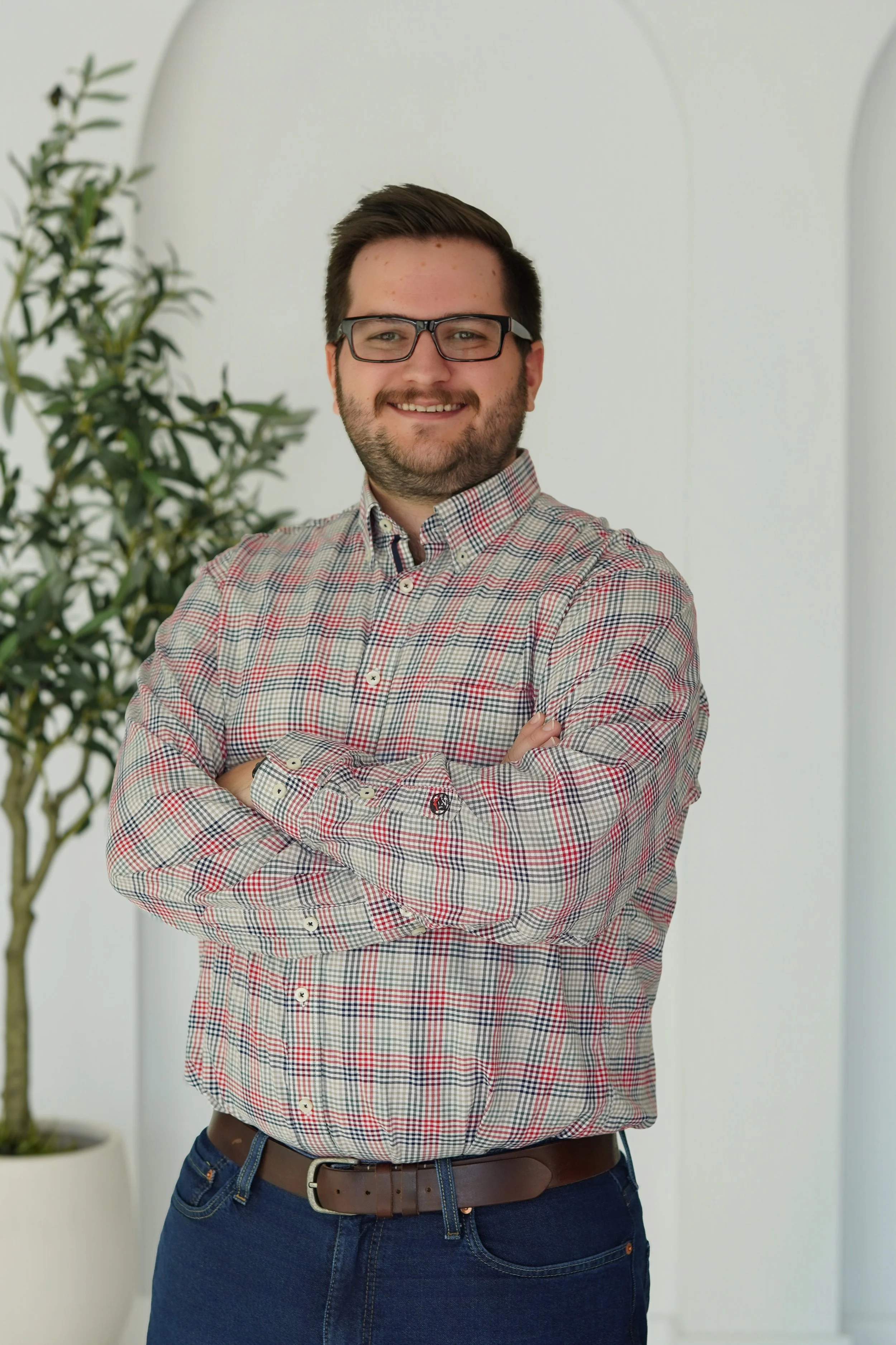 A young man with dark hair, beard, and glasses smiling with arms crossed, wearing a plaid shirt and blue jeans, standing in front of a white background with a potted plant.