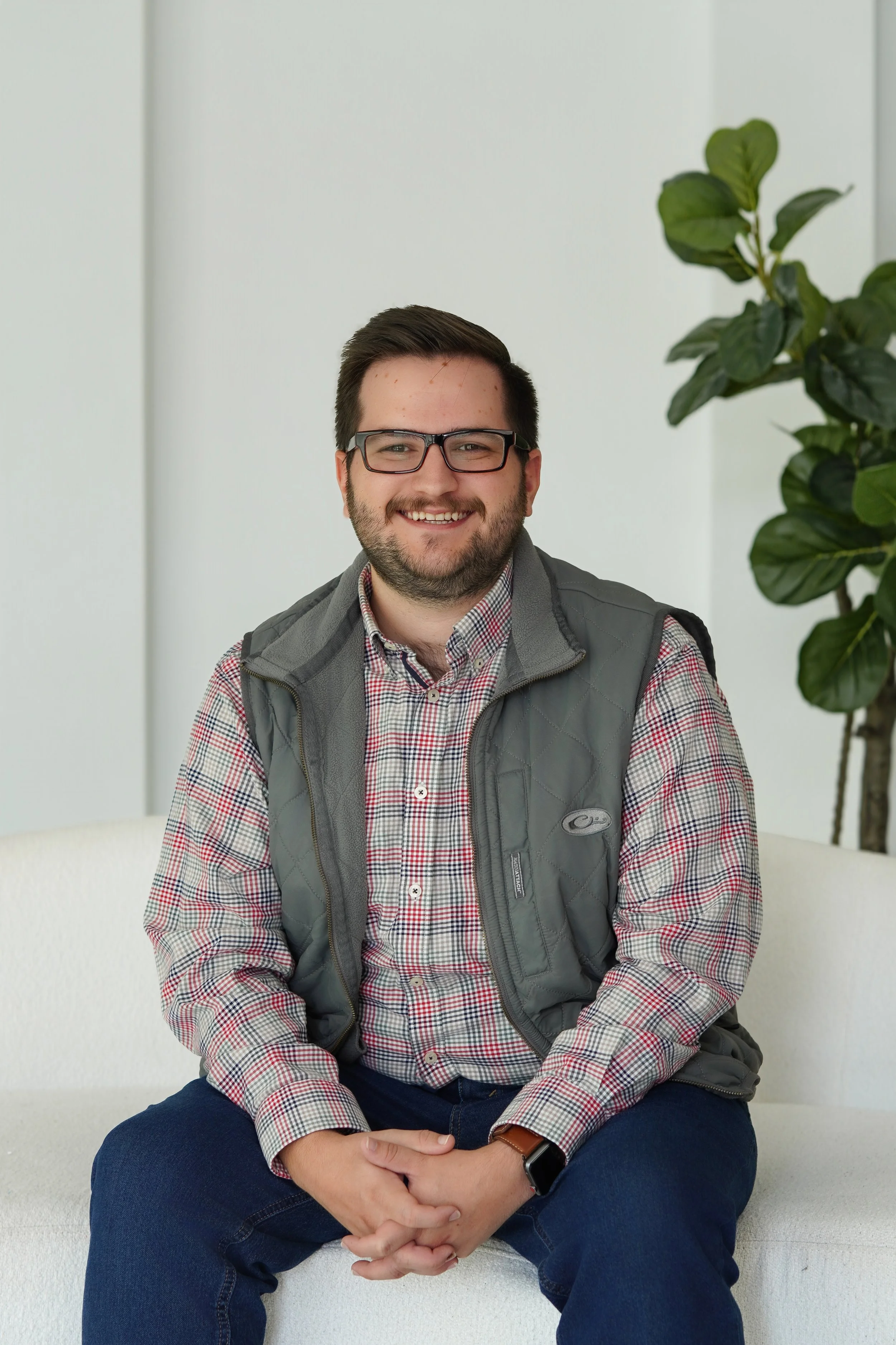 A man with short dark hair, glasses, and a beard, smiling and sitting on a white bench in front of a plain light-colored wall with a large green houseplant on his right side.
