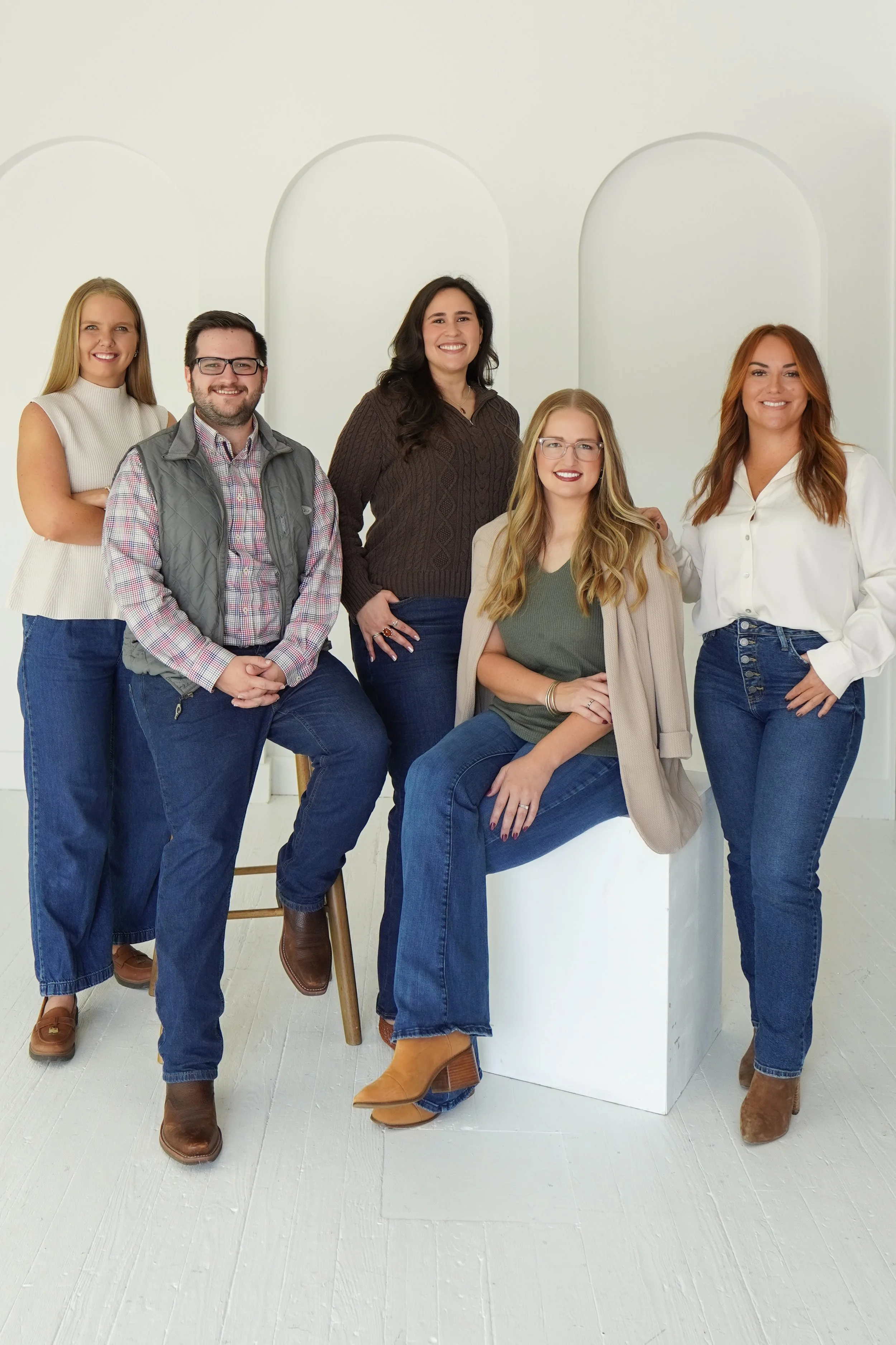 Group of six women posing together in a bright, white studio space, some standing and one sitting on a white block, all smiling and dressed in casual, stylish clothing with jeans and boots.