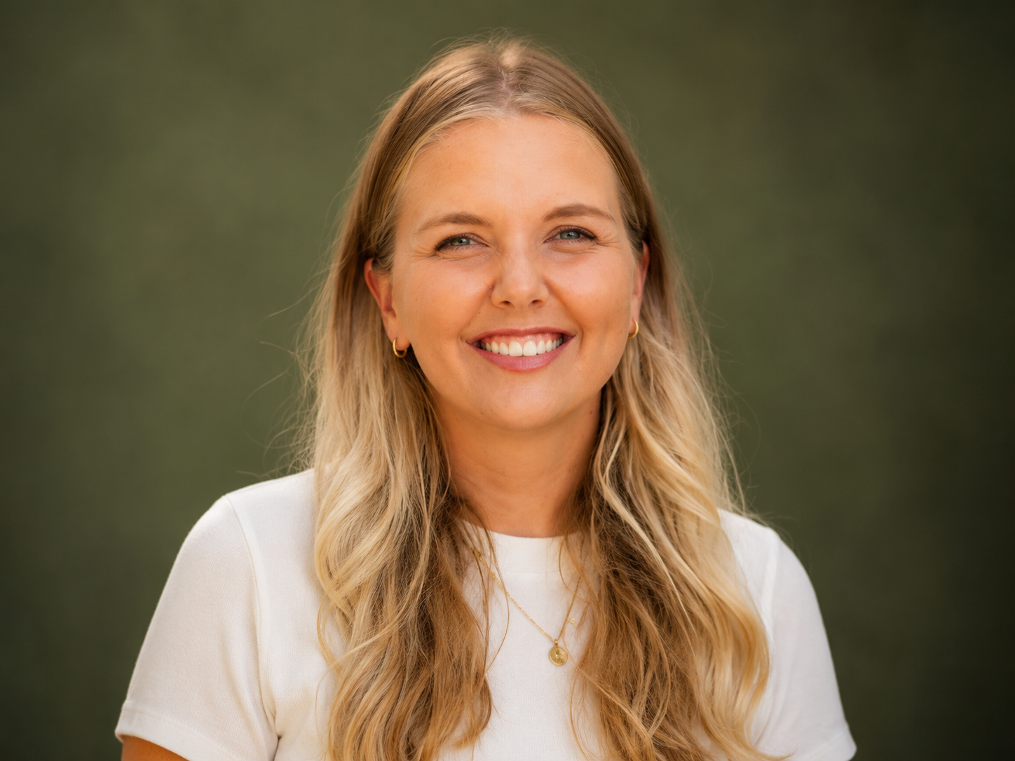 A smiling young woman with long blonde hair wearing a white shirt and gold jewelry standing outdoors with blurred green background.