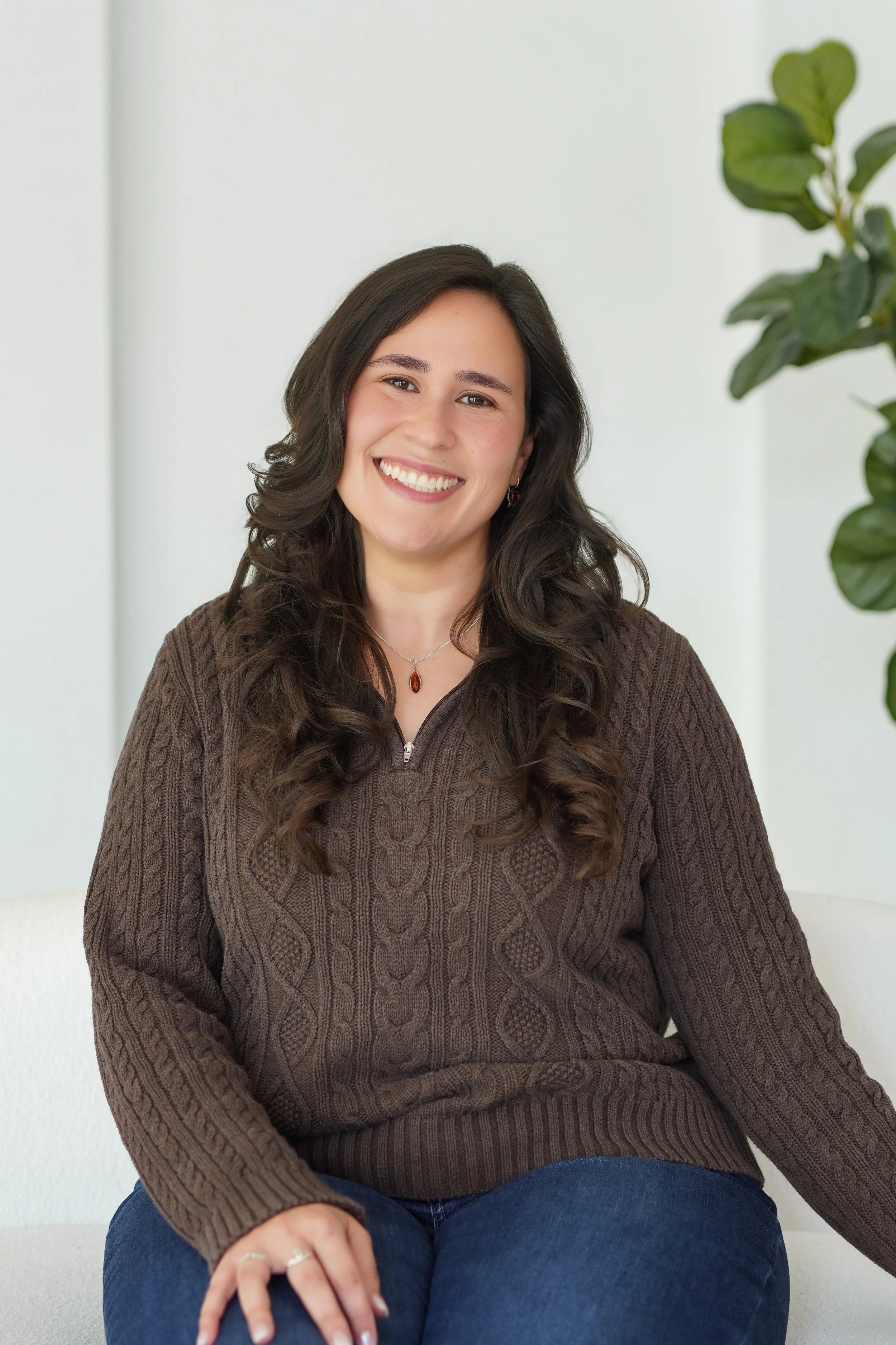A smiling woman with long dark hair wearing a brown sweater and blue jeans, sitting in front of a light-colored wall with a green plant on her right.