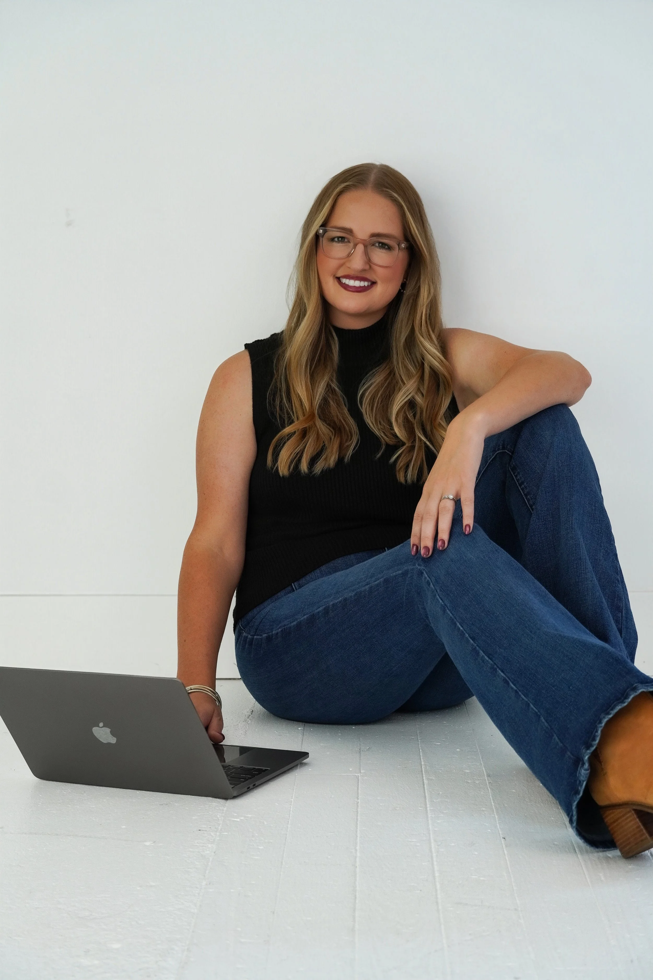 Woman with long hair, glasses, wearing a black sleeveless top and jeans, sitting on the floor with one knee up, smiling, and working on a MacBook laptop placed on a white surface against a white wall.