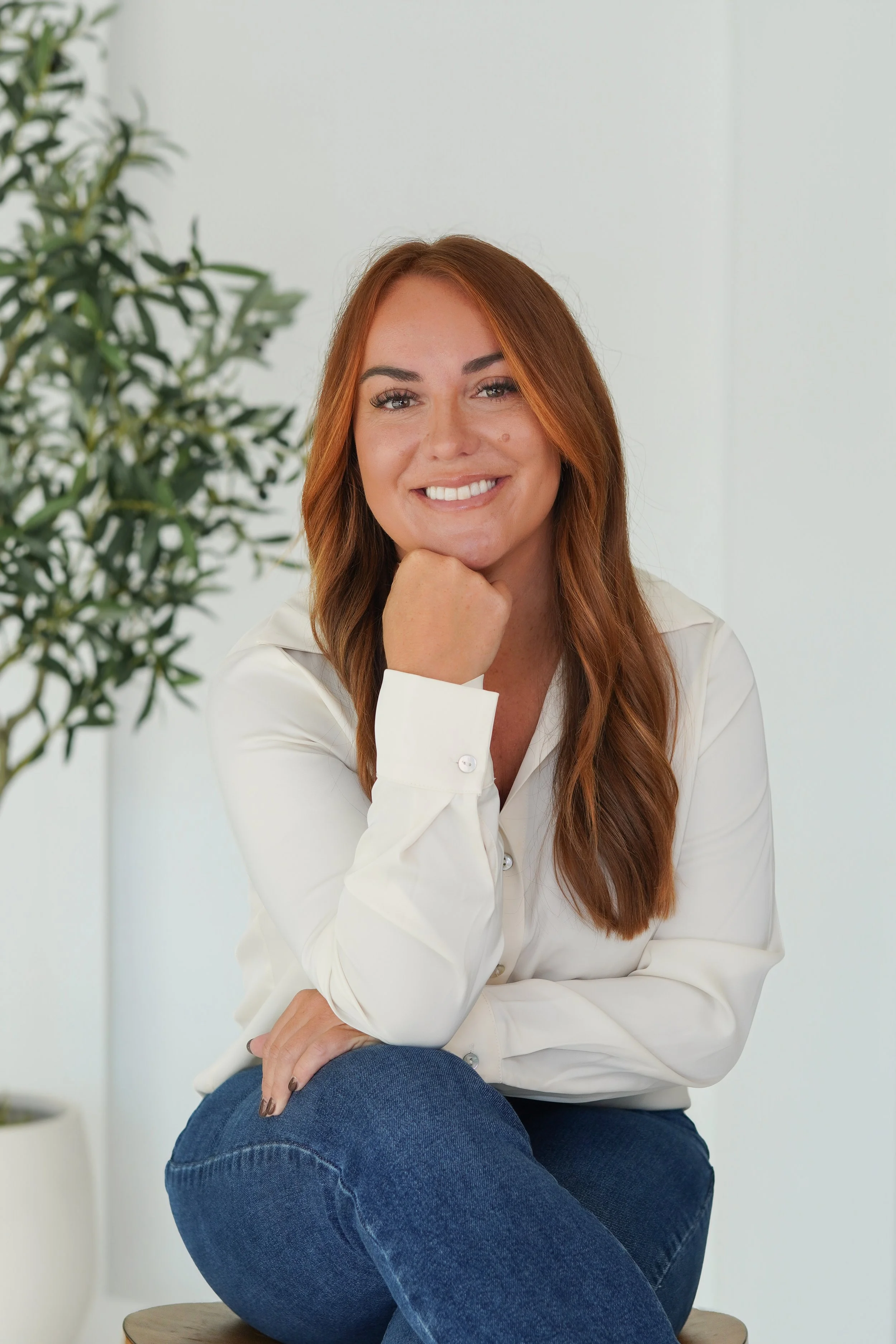 A woman with long red hair, wearing a white blouse, sitting on a stool in a minimalistic room with a plant in the background, smiling at the camera.