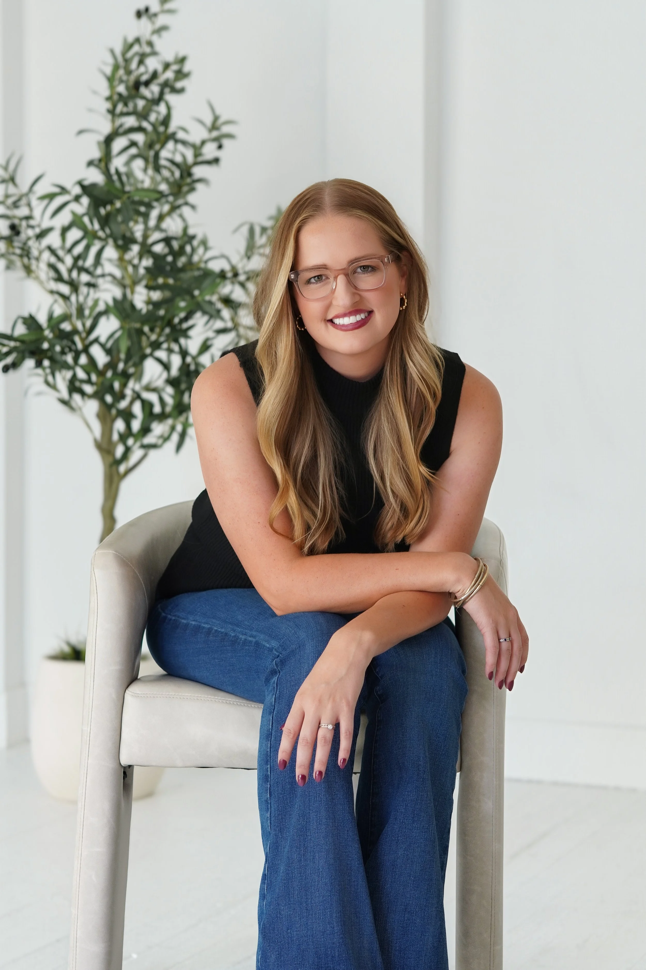 A smiling woman with long wavy blonde hair, wearing glasses, gold earrings, a black sleeveless top, and blue jeans, sits on a white chair with her arms resting on her knee in a bright, minimalist room with a potted plant in the background.