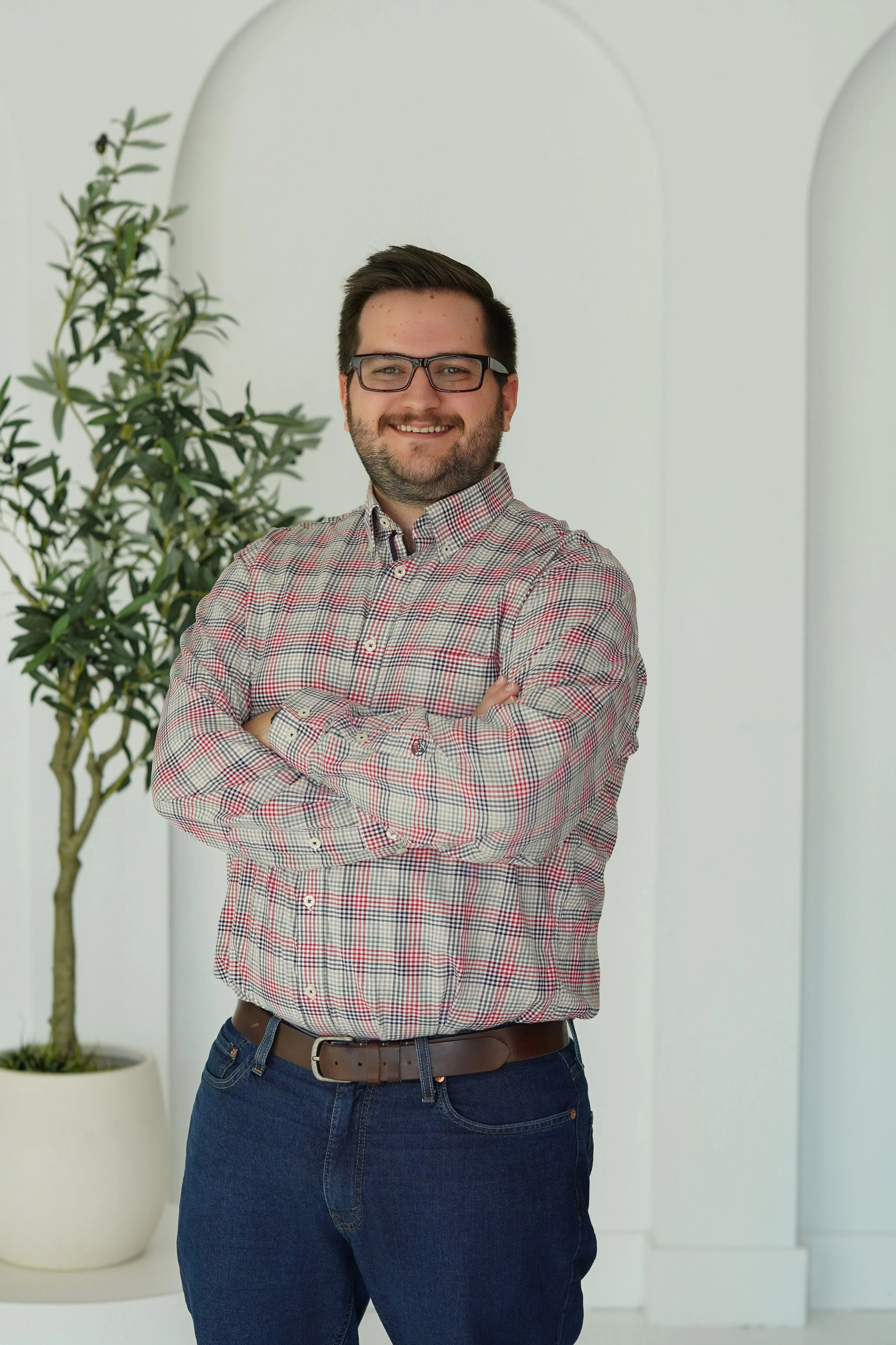 A man with glasses, a beard, and short dark hair smiling with arms crossed, wearing a plaid shirt and jeans, standing indoors next to a potted plant with green leaves.