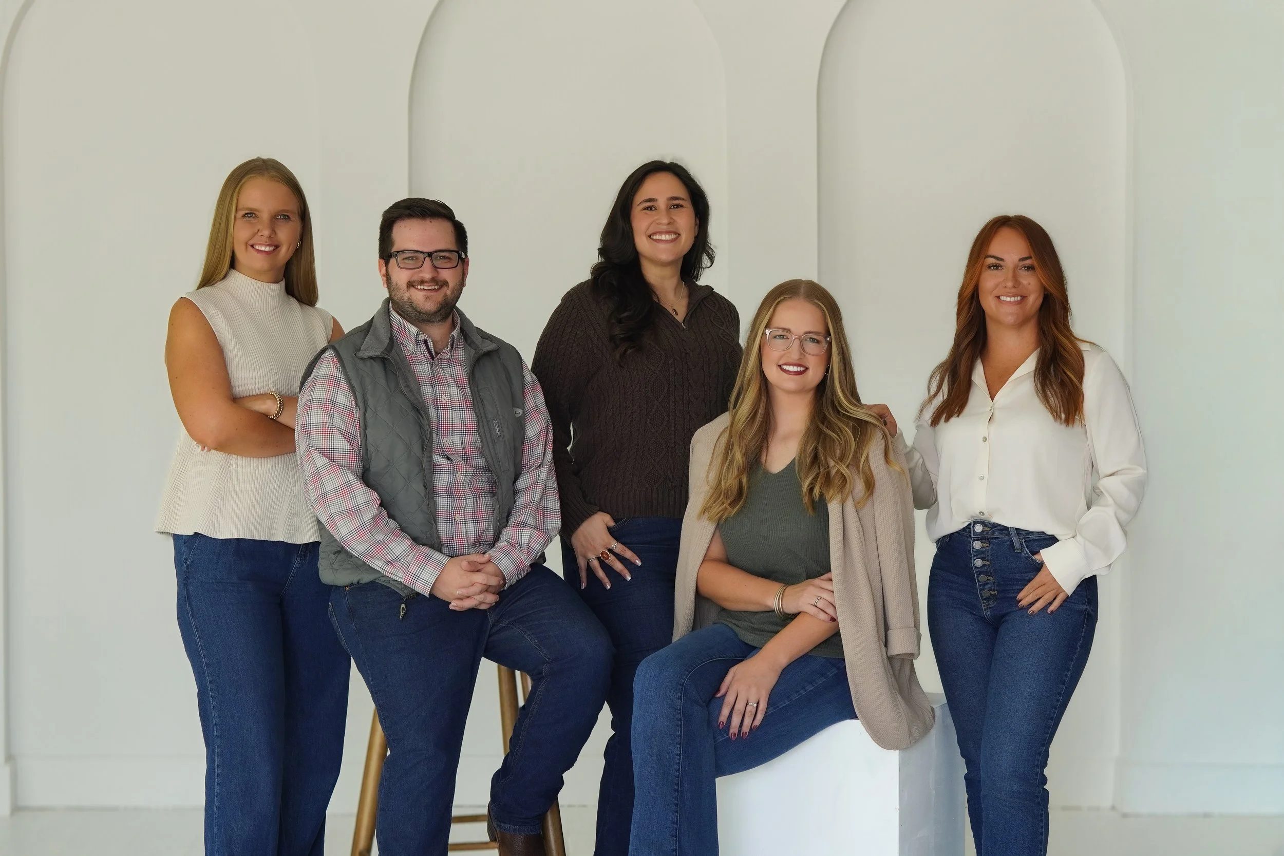 Group of five diverse women and one man posing together in front of a plain white wall, smiling.