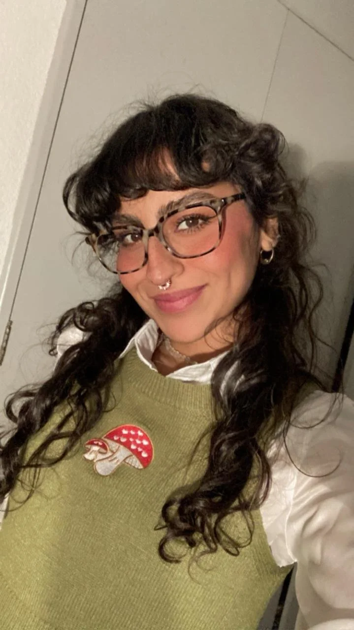 Joven con gafas, cabello rizado y peinado con flequillo, usa una blusa blanca y un suéter verde con un parche de hongo en el pecho, sonriente frente a una pared clara.