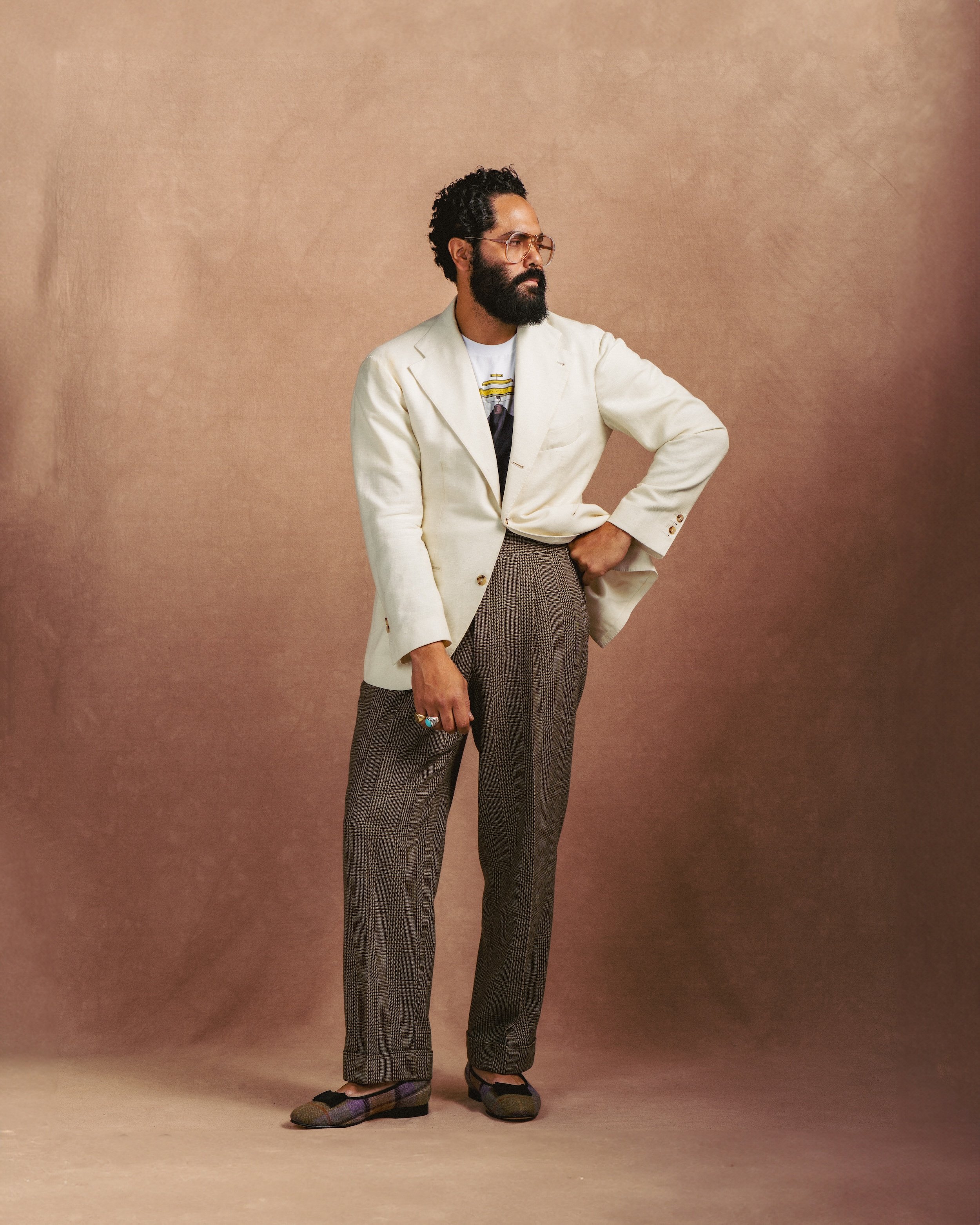 Man in stylish attire posing against a plain background, wearing a cream blazer, a t-shirt, plaid trousers, and loafers, with glasses and a beard.