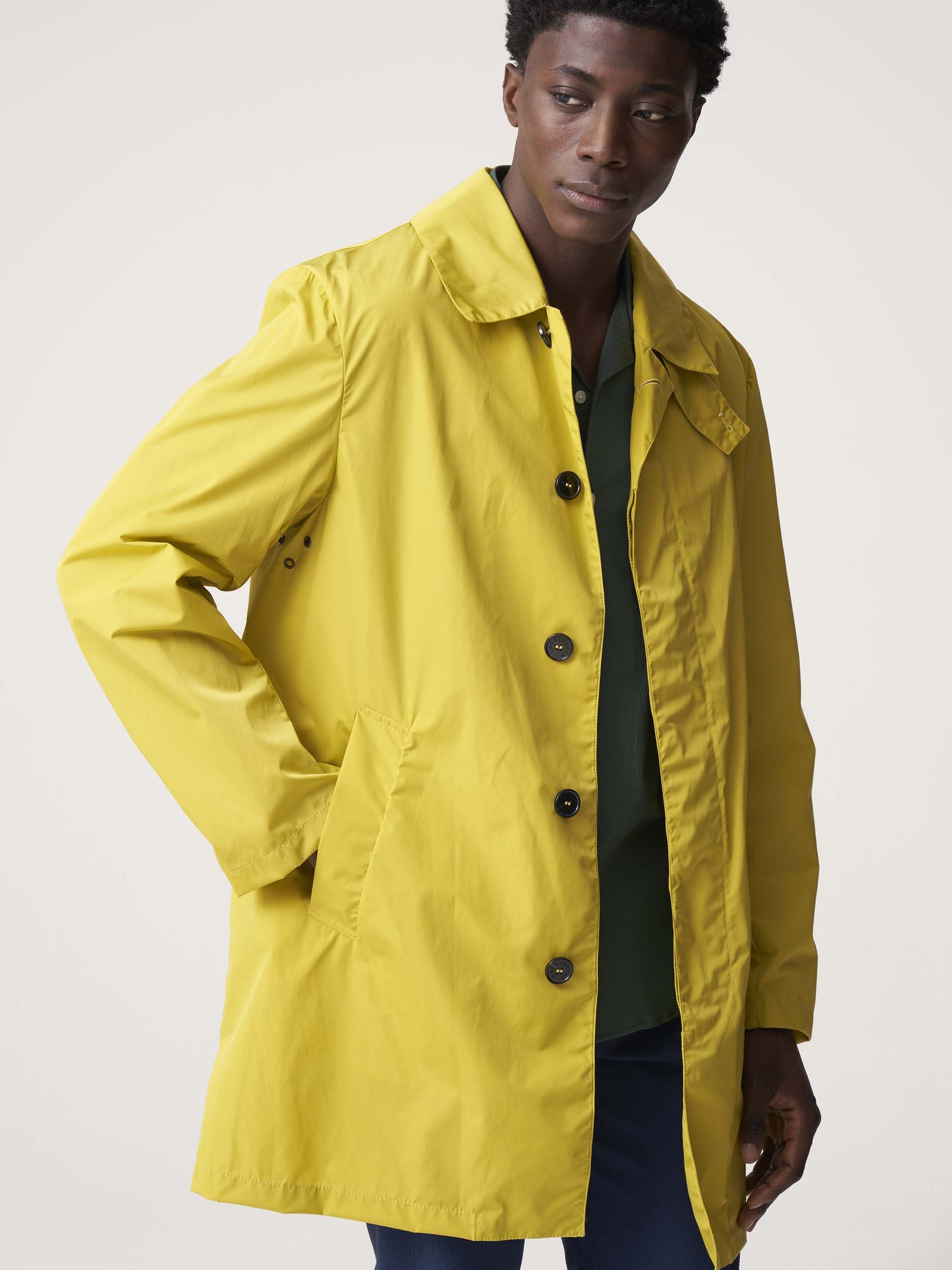 Man wearing a yellow trench coat with a green shirt underneath, posing against a plain background.