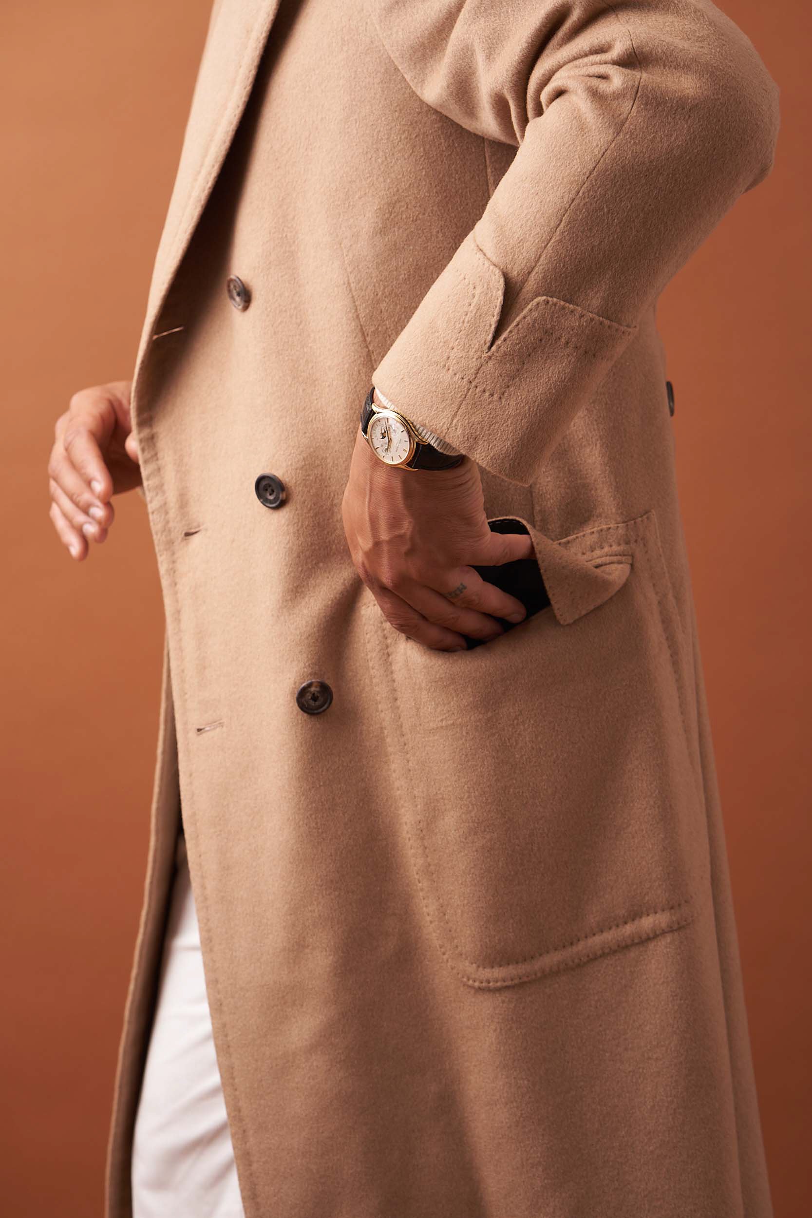 Person wearing a beige overcoat with a watch, hand in pocket.