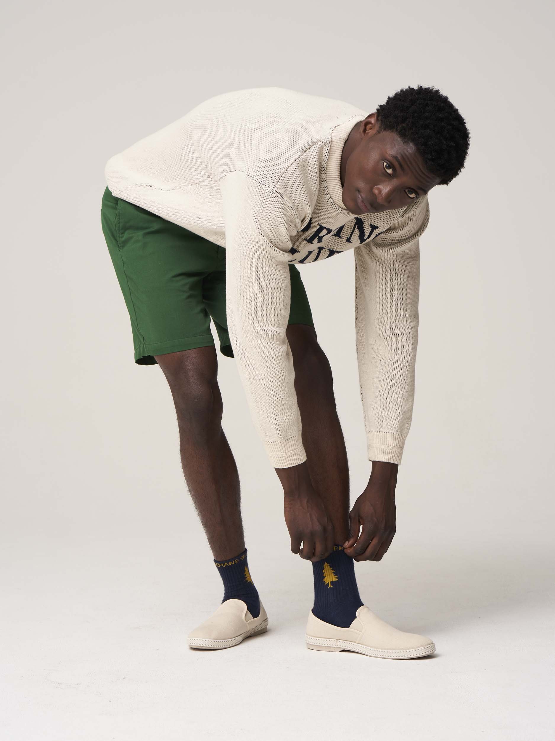 Man in casual clothing adjusting socks, wearing green shorts, a cream sweater, and cream slip-on shoes.