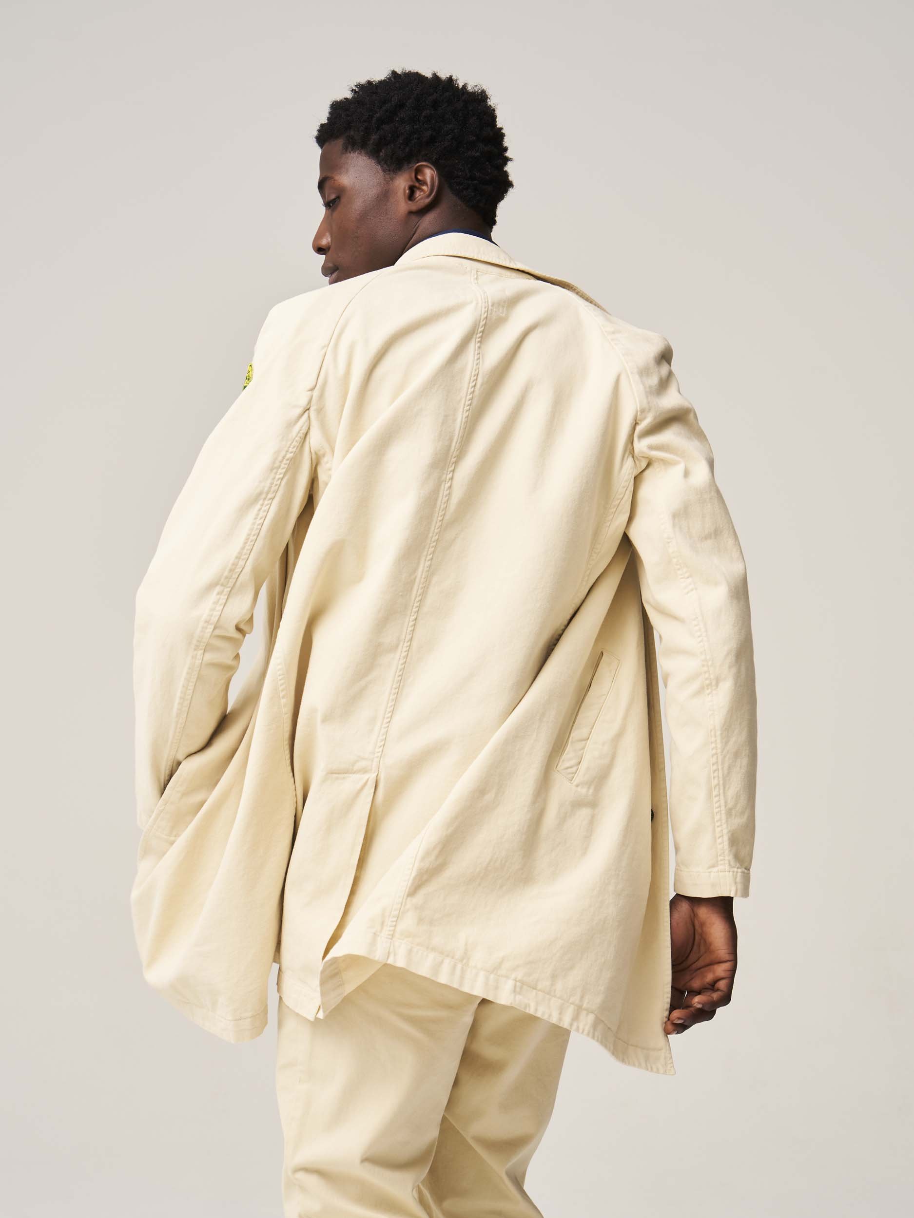Man wearing beige jacket and pants, back view, against a light background.