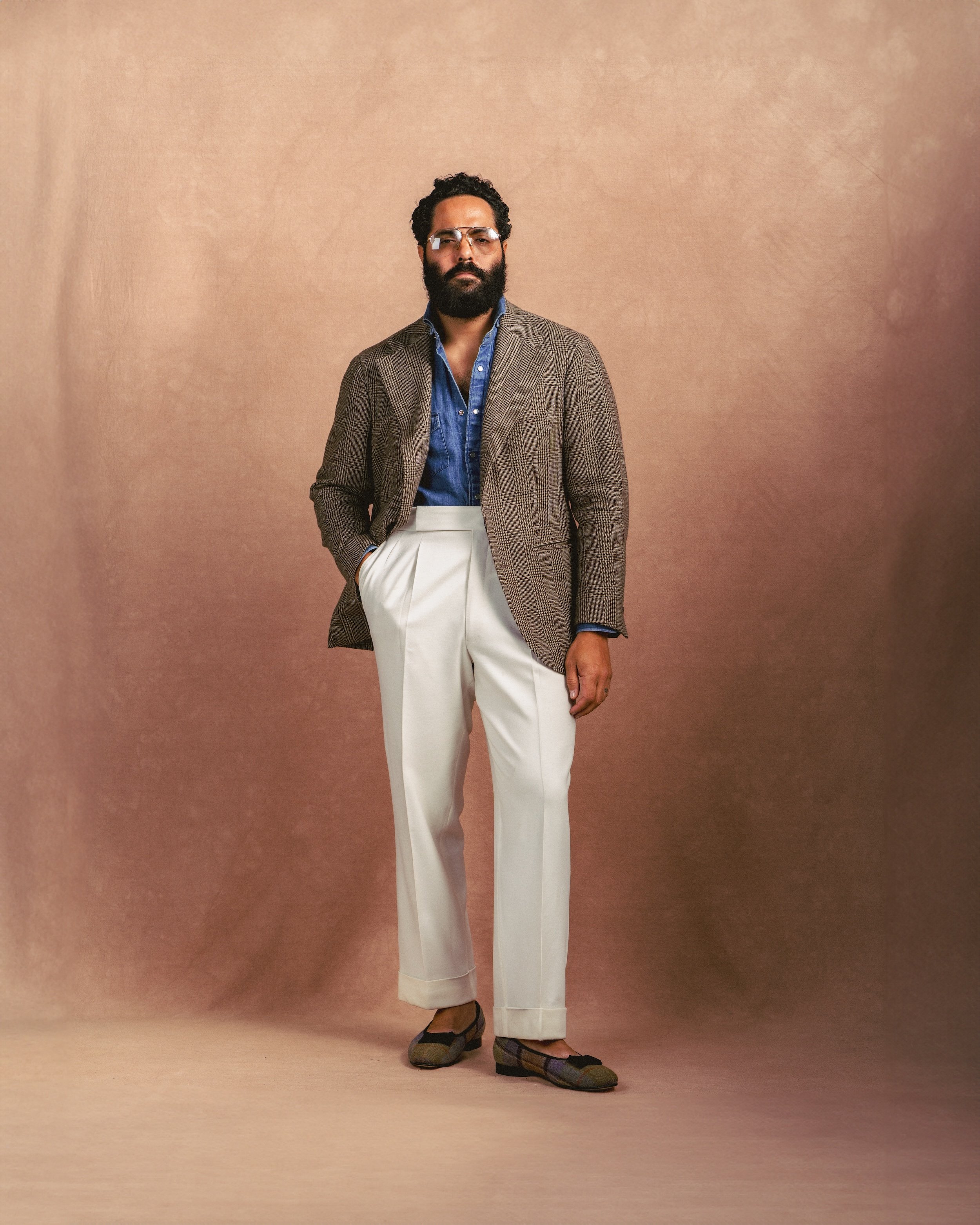 Man in stylish outfit with blazer, blue shirt, white pants, and loafers standing against a brown backdrop.