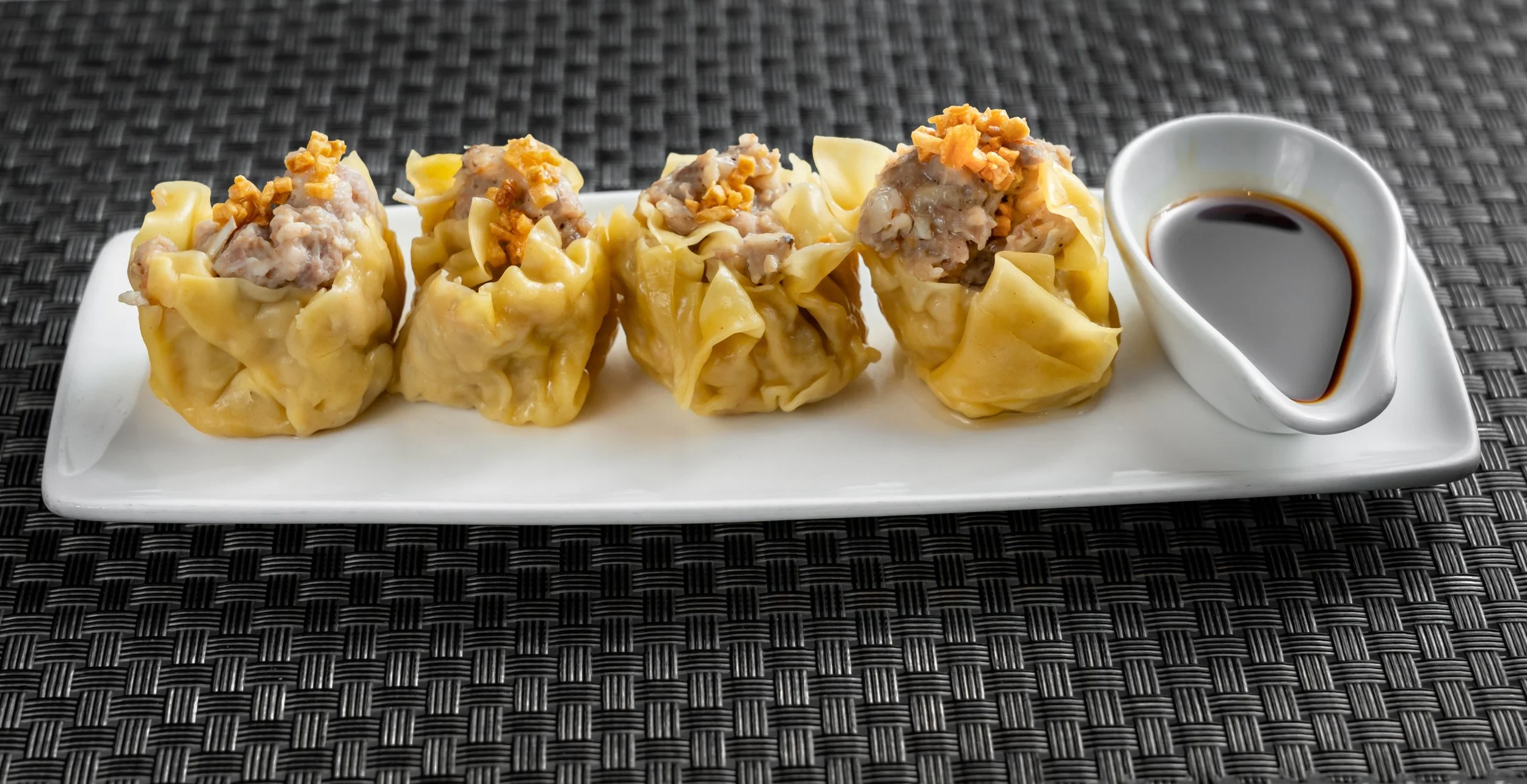 Plate of shumai dumplings topped with crispy garlic and served with soy sauce on the side, placed on a woven mat.