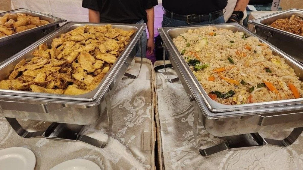 Buffet trays with seasoned grilled chicken and vegetable fried rice on a table with a white tablecloth.