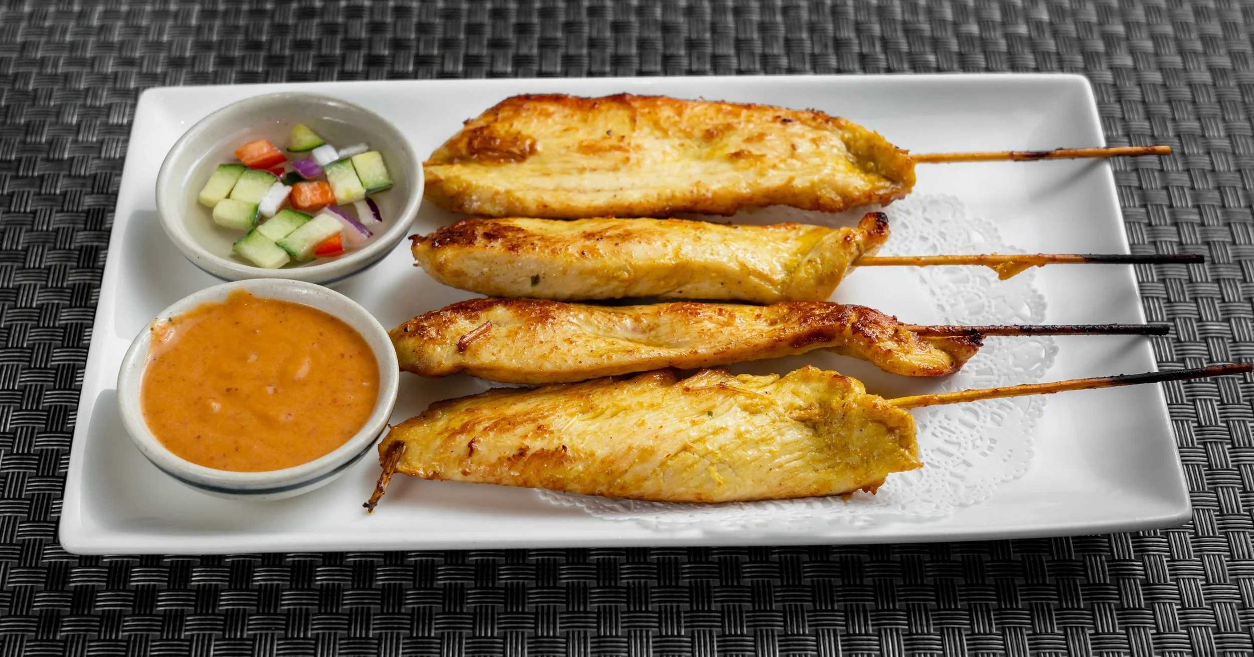 Grilled chicken skewers served on a rectangular plate with peanut sauce and a side of mixed cucumber, tomato, and red onion salad.