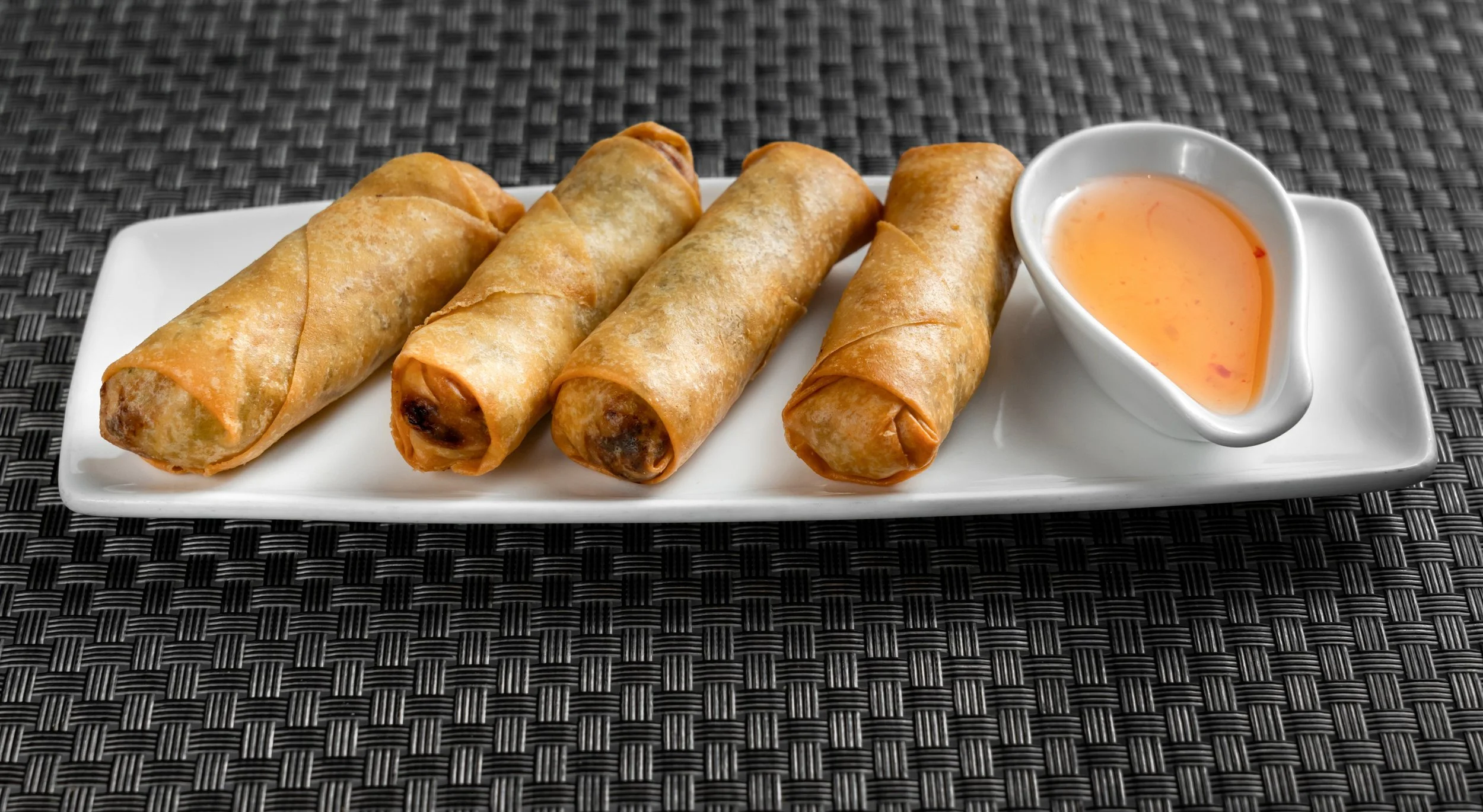 Four spring rolls on a white plate with dipping sauce on a woven placemat.