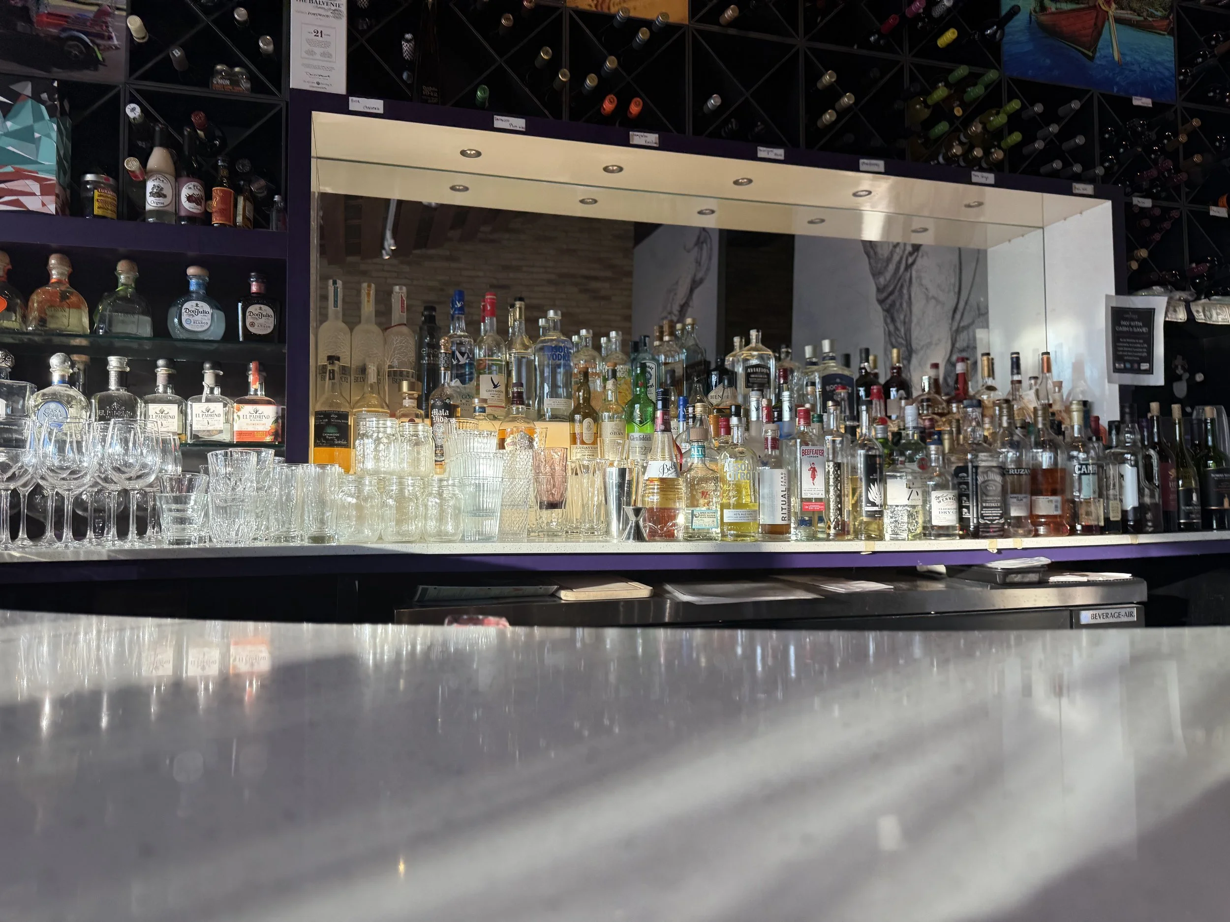 Bar counter with various liquor bottles, wine glasses, and shelves displaying more bottles in the background.