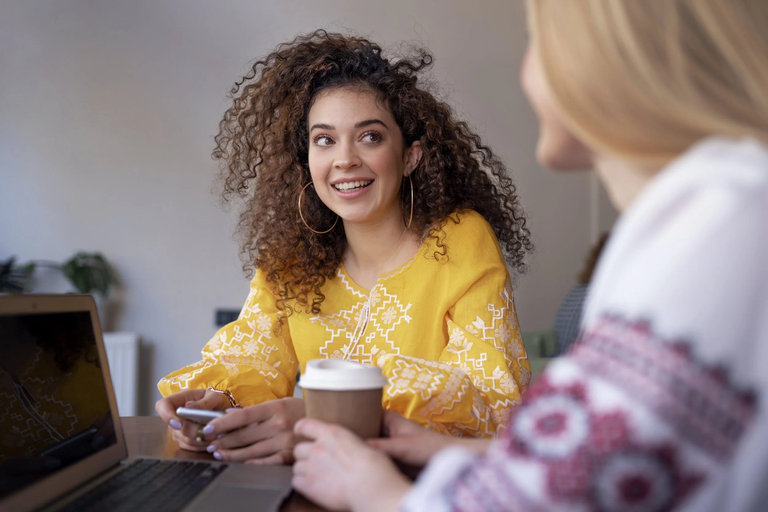Two women sitting at a table in conversation, with a laptop and coffee cup nearby, suggesting a calm one-to-one support discussion.