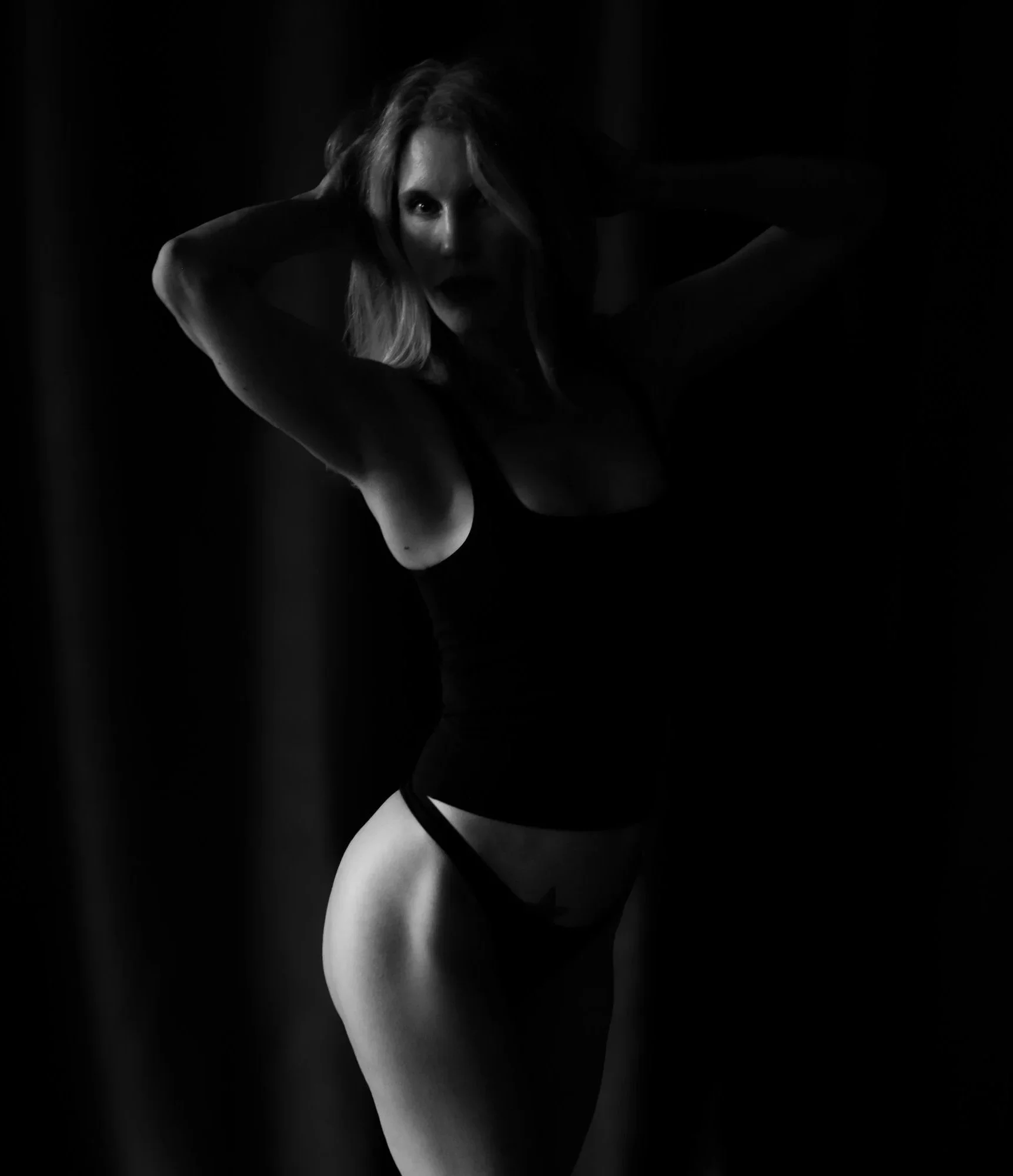 A woman posing in black lingerie in a dark setting. She has her hands in her hair and is looking towards the camera.