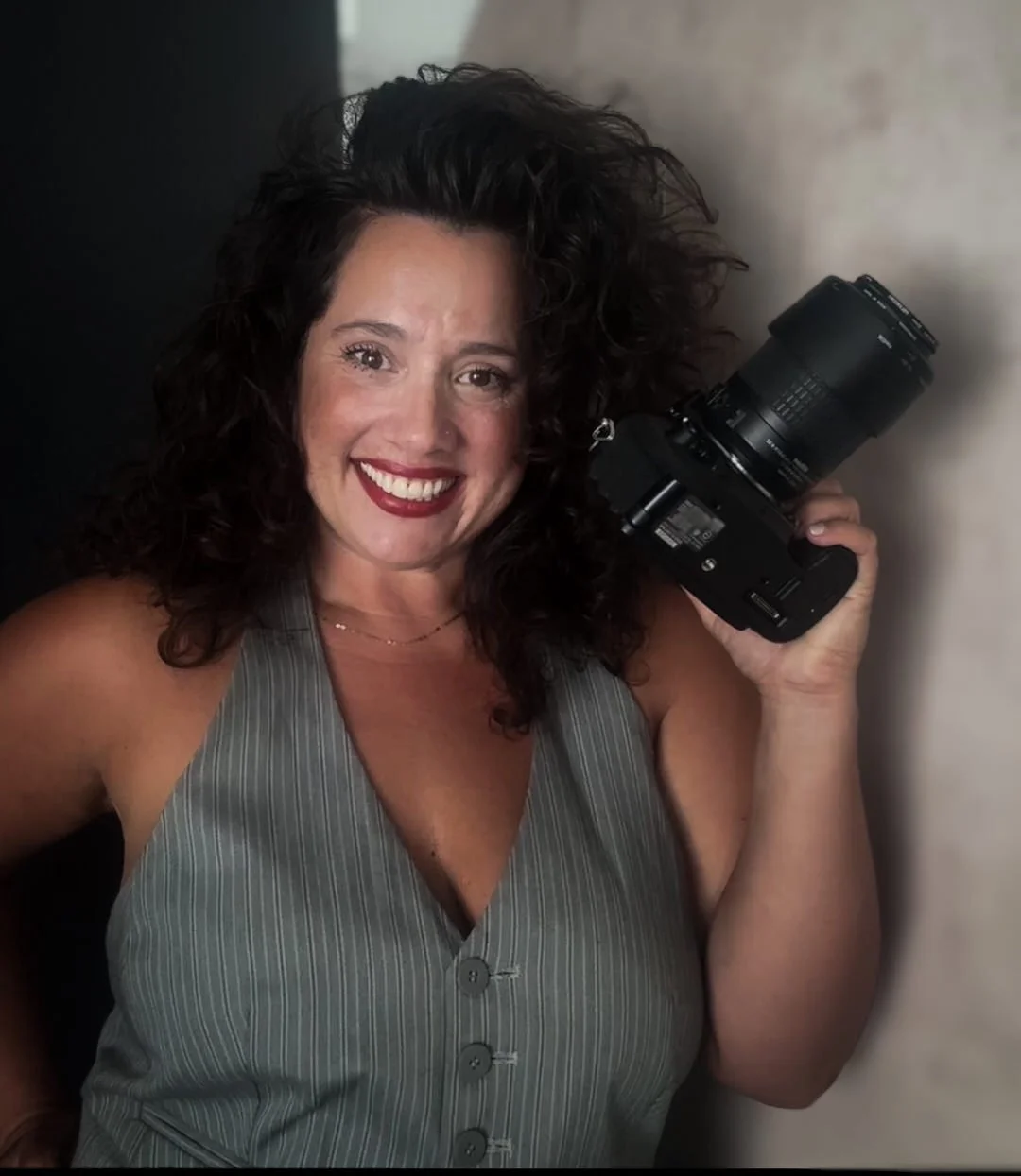 A woman with curly dark hair, smiling, holding a camera in her right hand, wearing a sleeveless striped gray top.