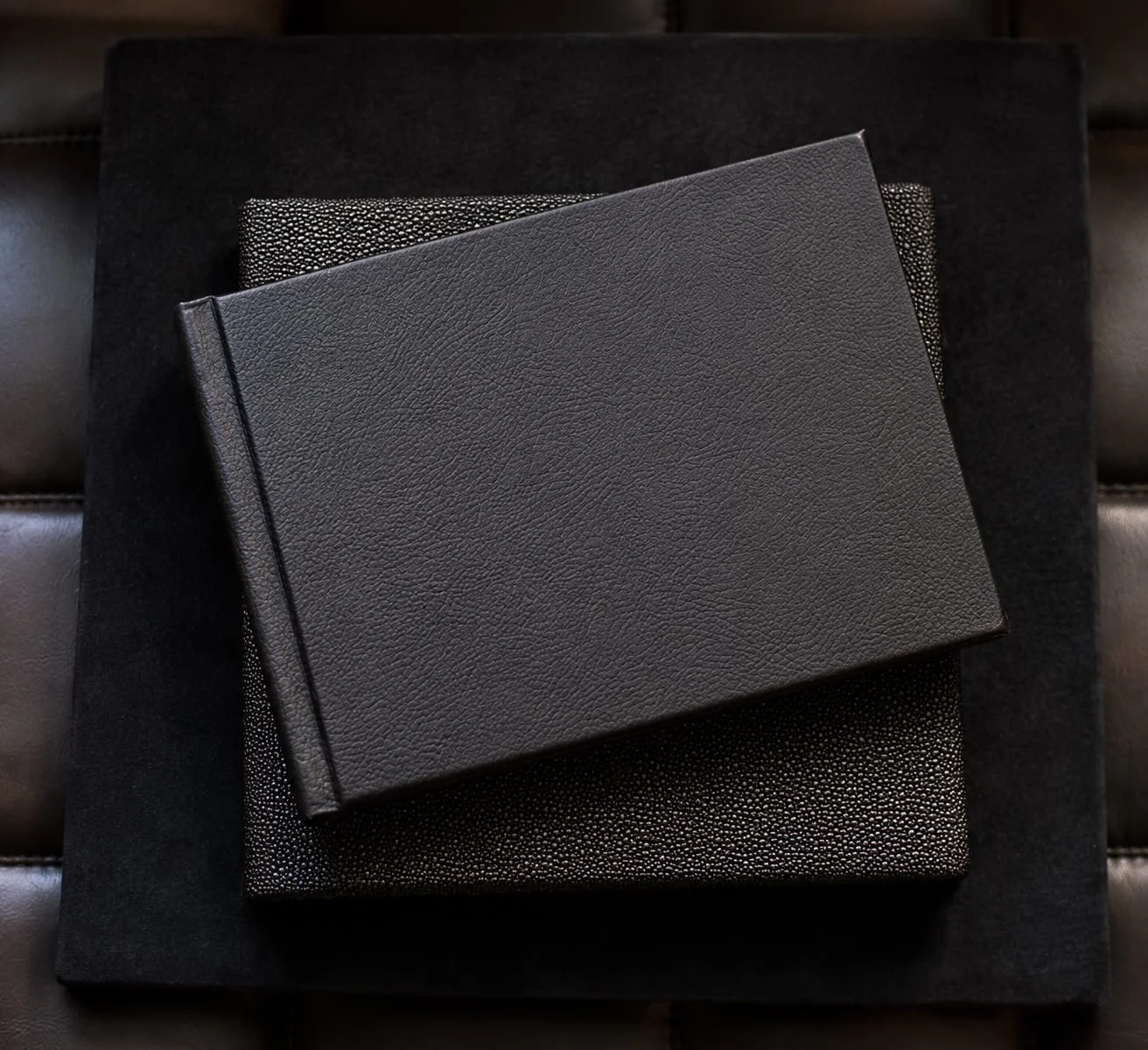 A black textured leather-bound book on top of two other black books, one with a spottier pattern and one with a smooth surface.
