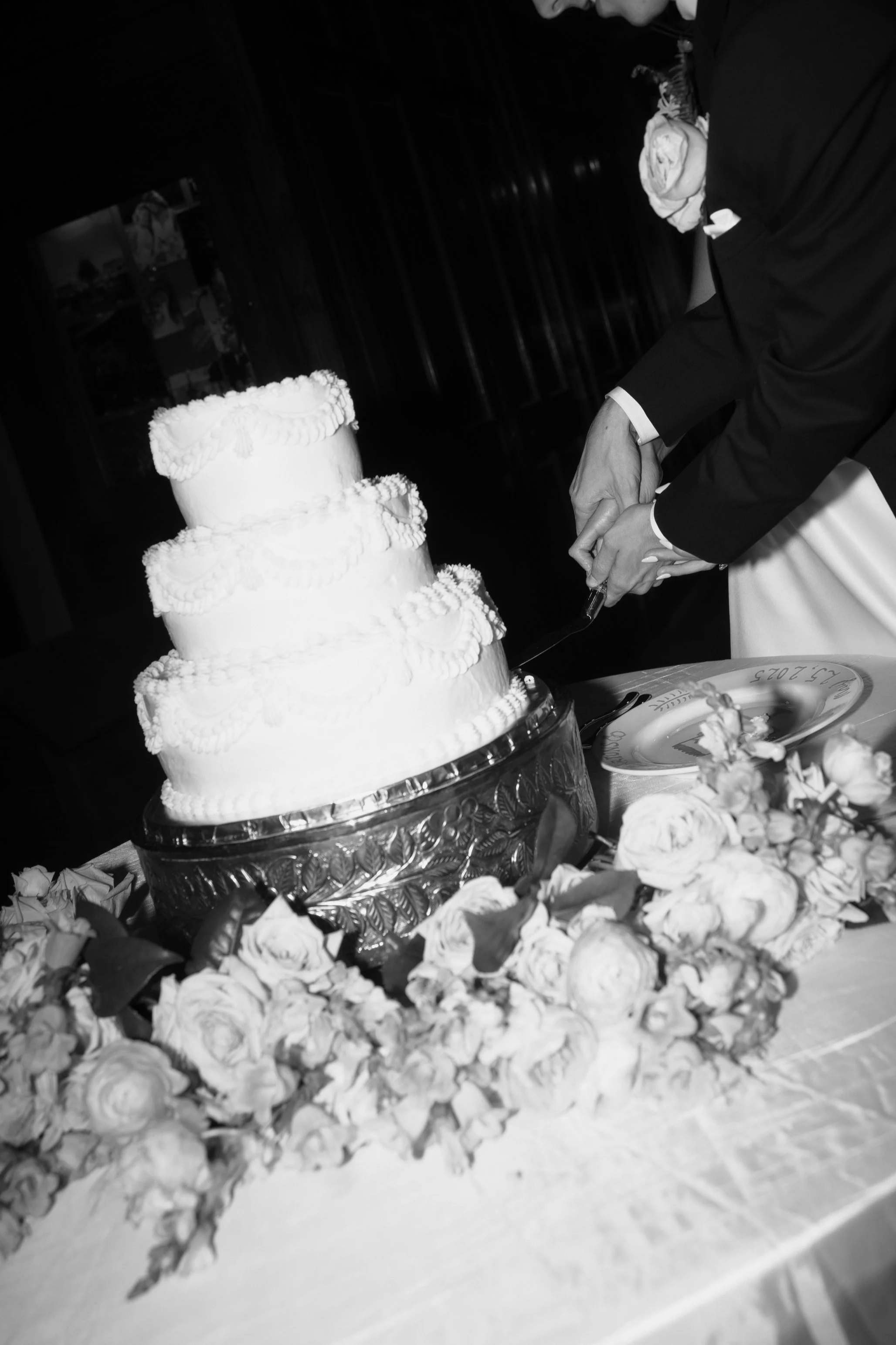 A groom in a tuxedo cuts a wedding cake decorated with white frosting, surrounded by a floral arrangement on a table.